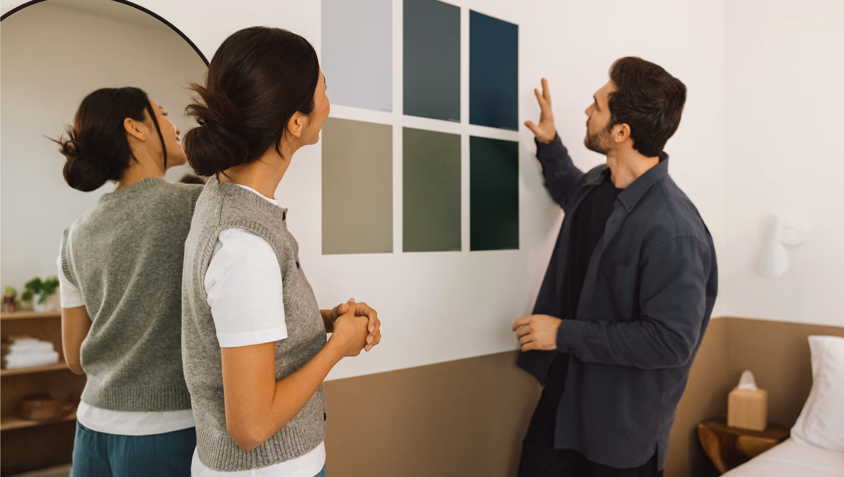 Man and woman standing indoors looking at six color swatches arranged in two rows on a wall.