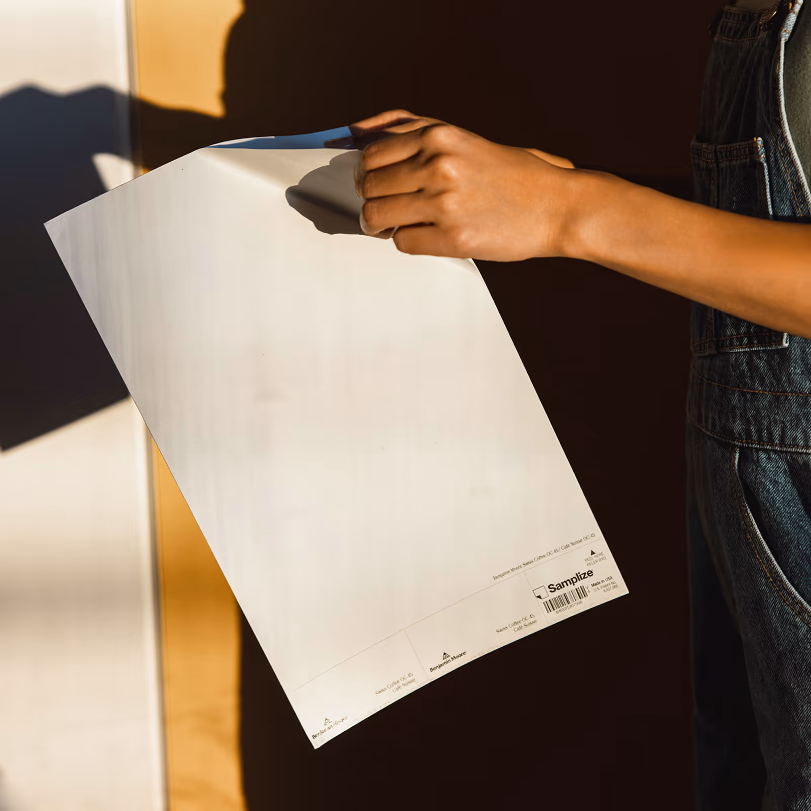 Person holding a white Benjamin Moore paint sample sheet with shadow on a dark background.