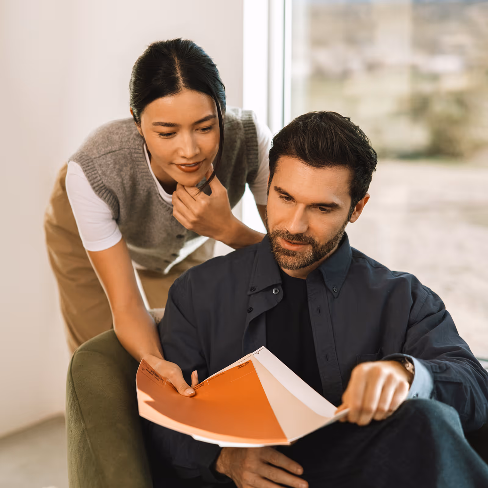 Man sitting in chair reading documents while woman leans over his shoulder, looking at papers together.
