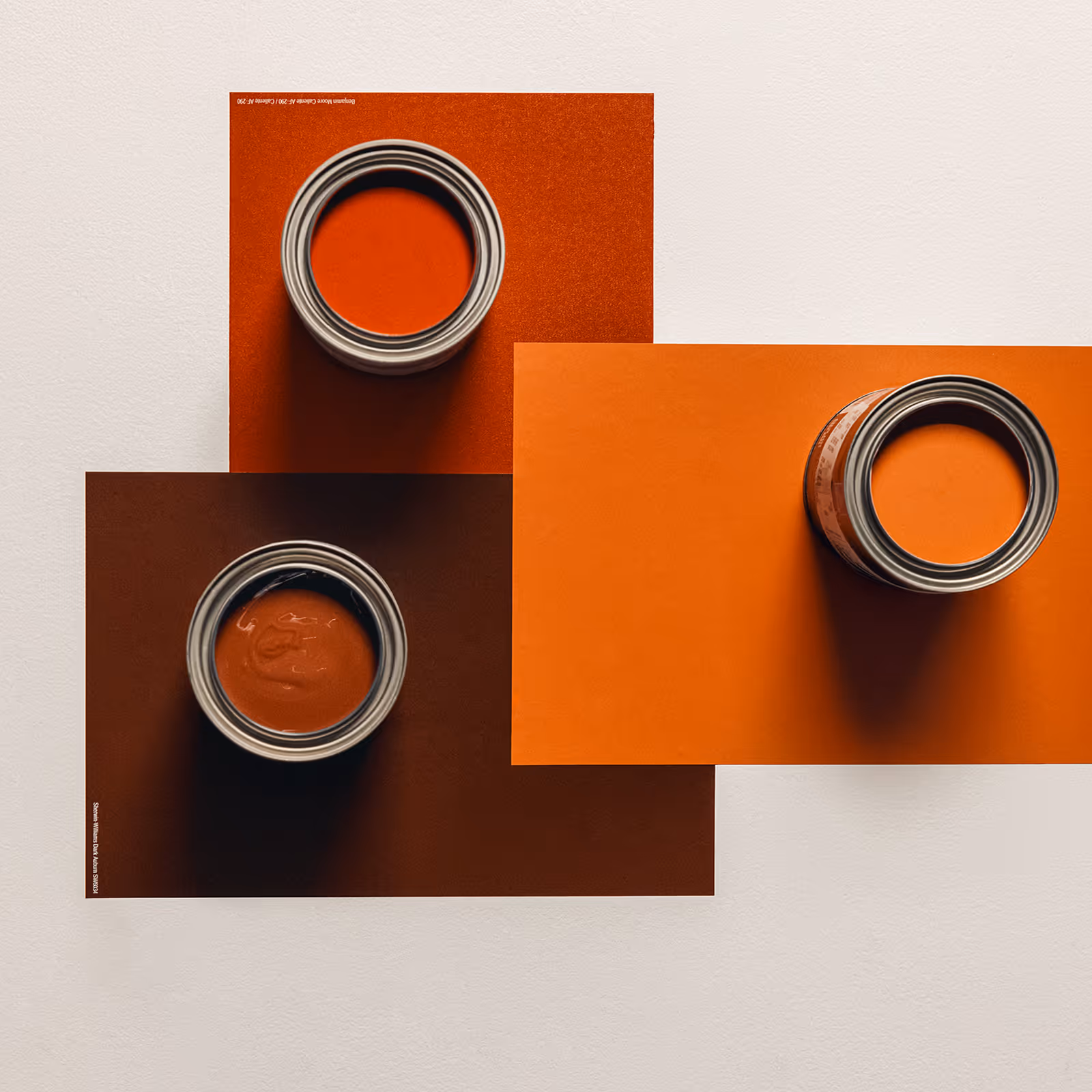 Three open paint cans with different shades of orange paint on rectangular color samples.