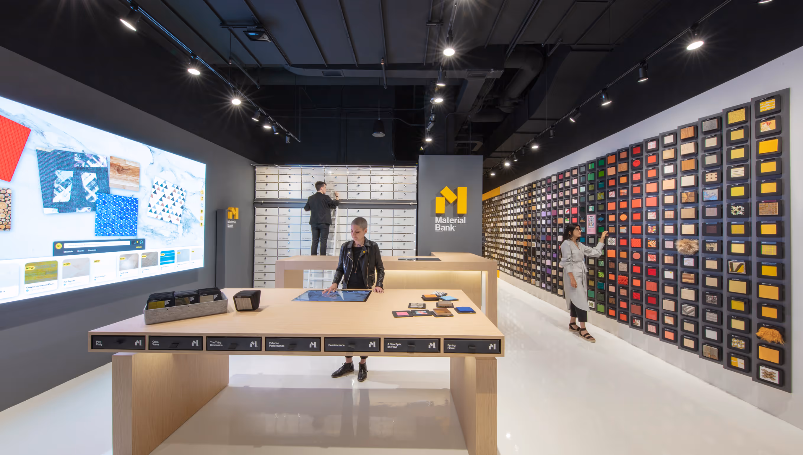 Modern showroom with a large touch table, wall-mounted material samples, and people interacting with displays.