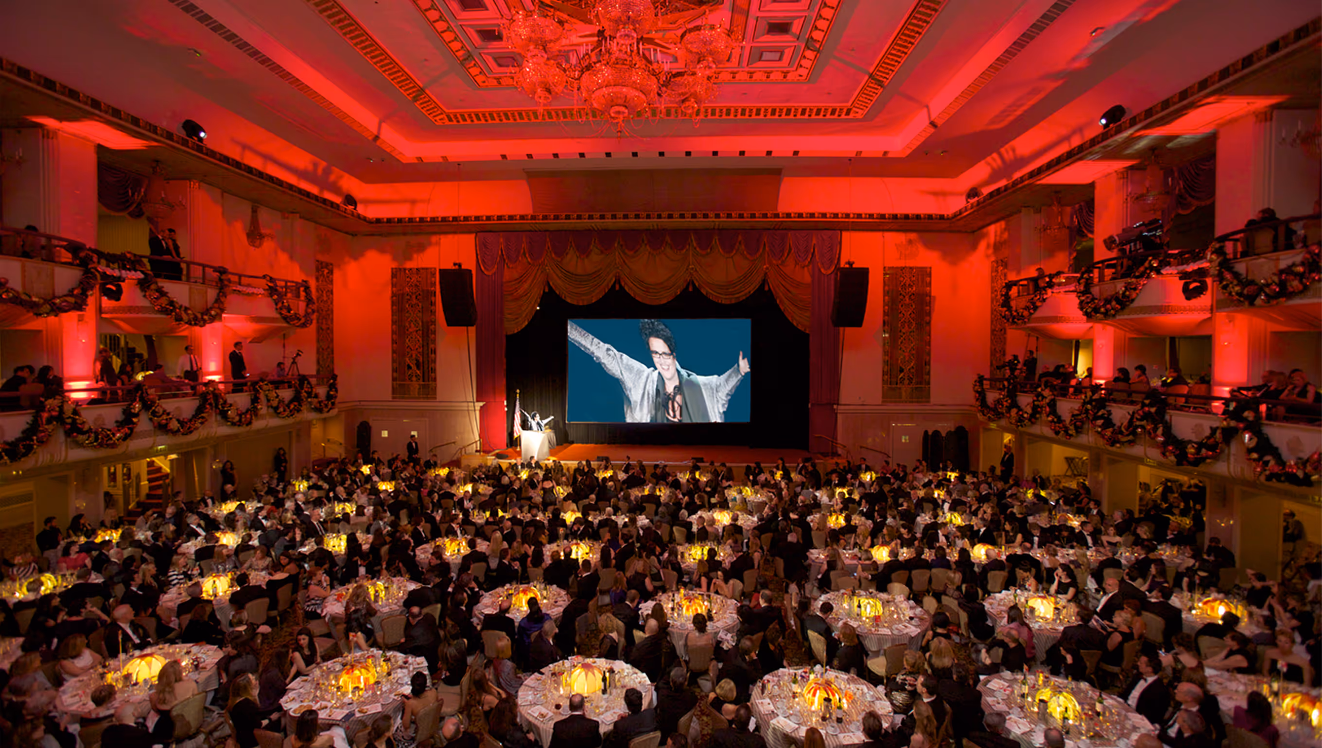 Large formal banquet with round tables and guests watching a speaker at a podium under red lighting in an ornate hall.