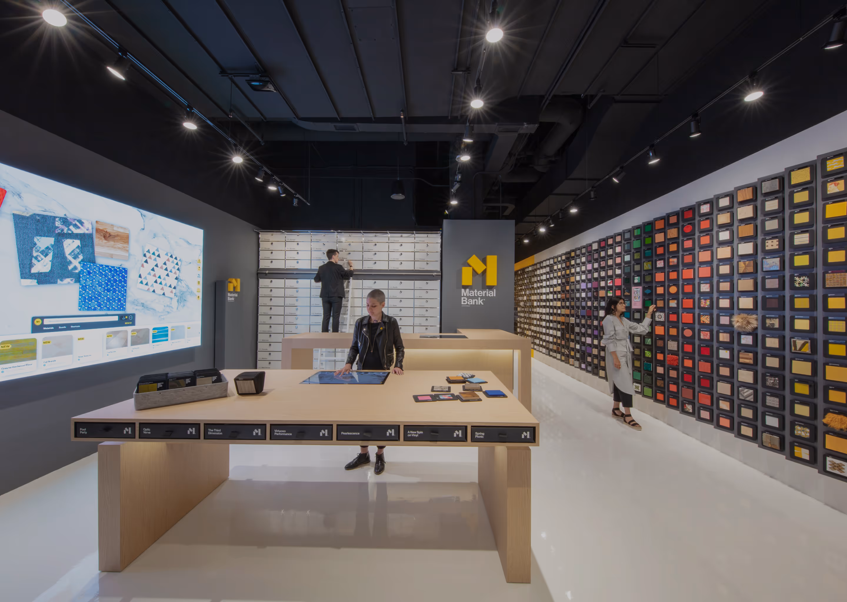Modern interior of Material Bank with sample walls, a woman using a touchscreen table, and another woman browsing material samples on a large colorful wall display.