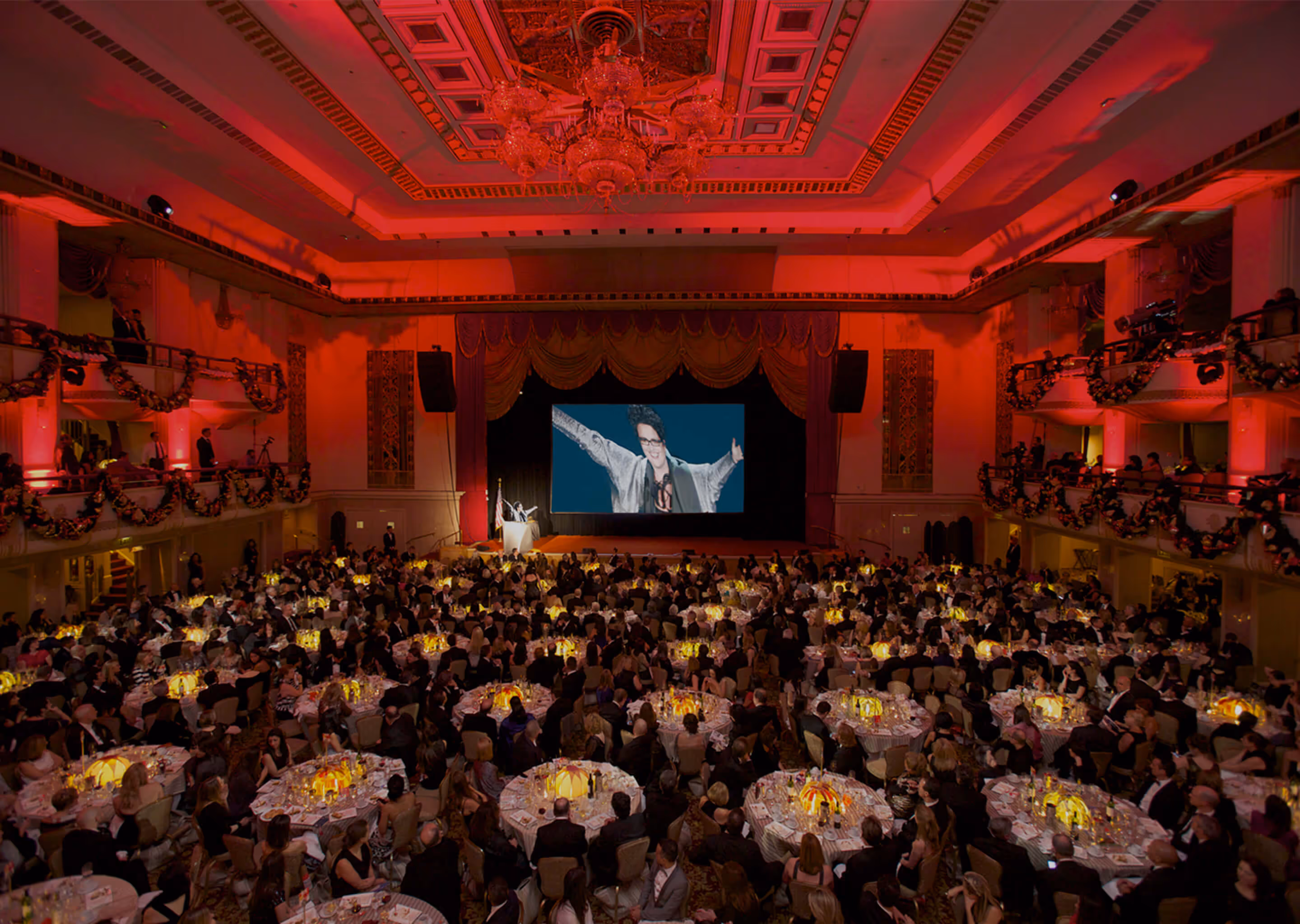 Large banquet hall with many round tables filled with guests watching a stage featuring a podium and a large screen showing a person with arms outstretched.