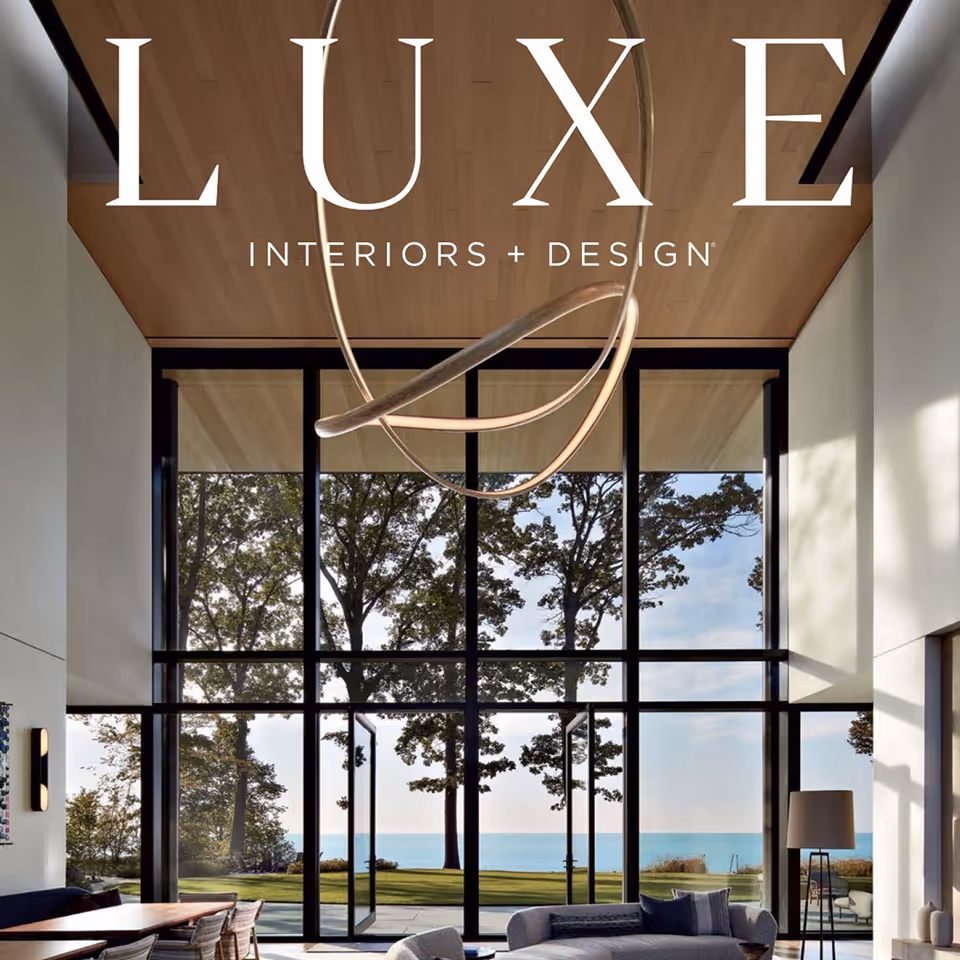 Modern living room with large floor-to-ceiling windows overlooking trees and a lake, featuring a curved sofa and wooden dining table.