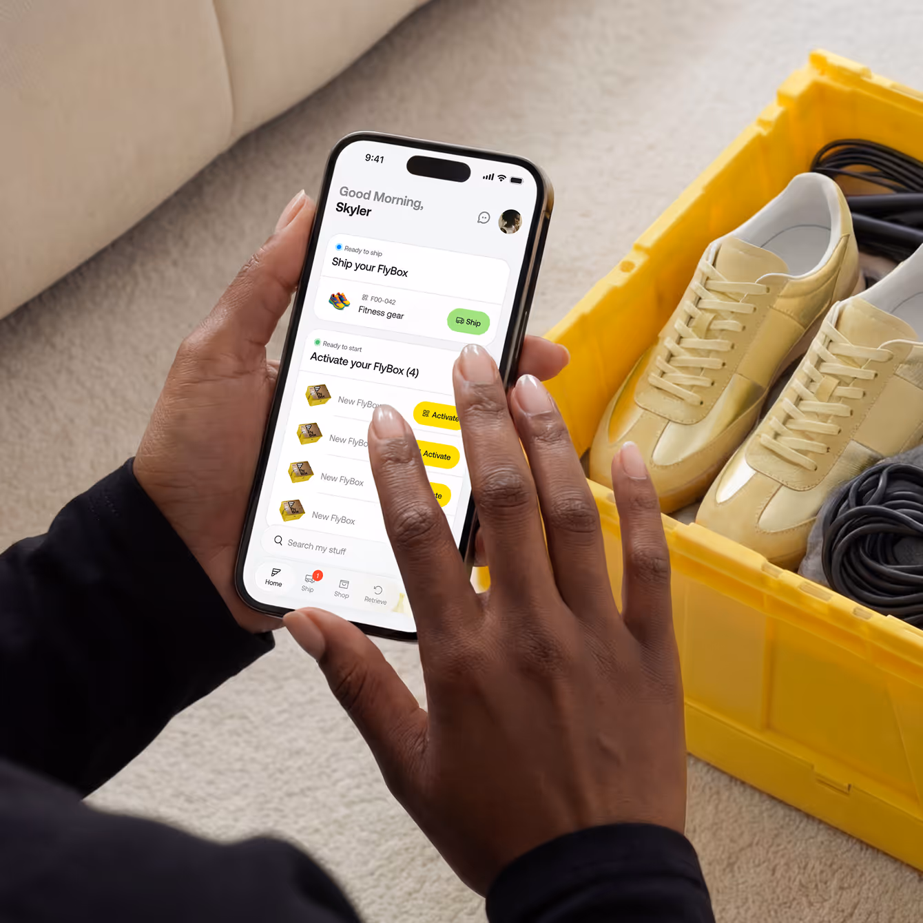Person holding a smartphone displaying an app with options to ship and activate FlyBoxes, next to a yellow crate containing yellow sneakers and cables.