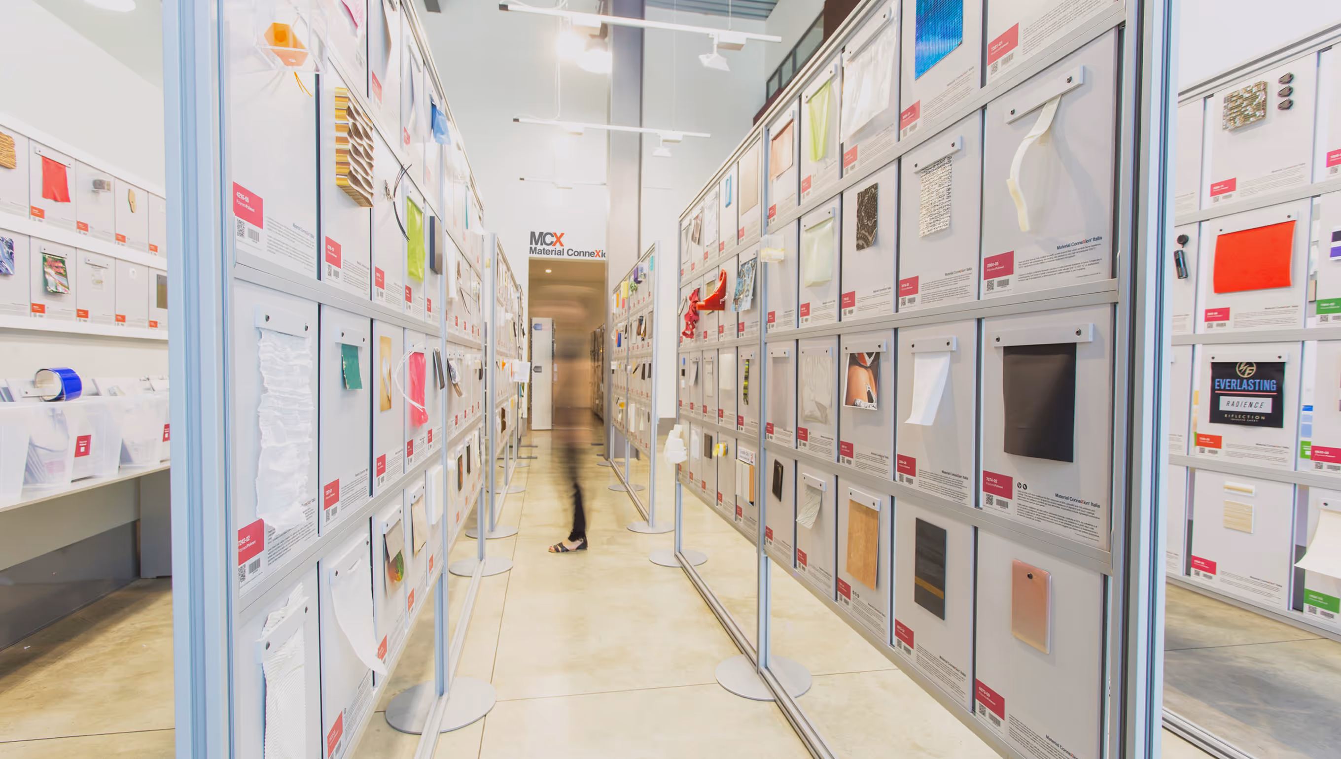 Indoor exhibition corridor with samples of materials displayed on panels along both sides and a blurred person walking.