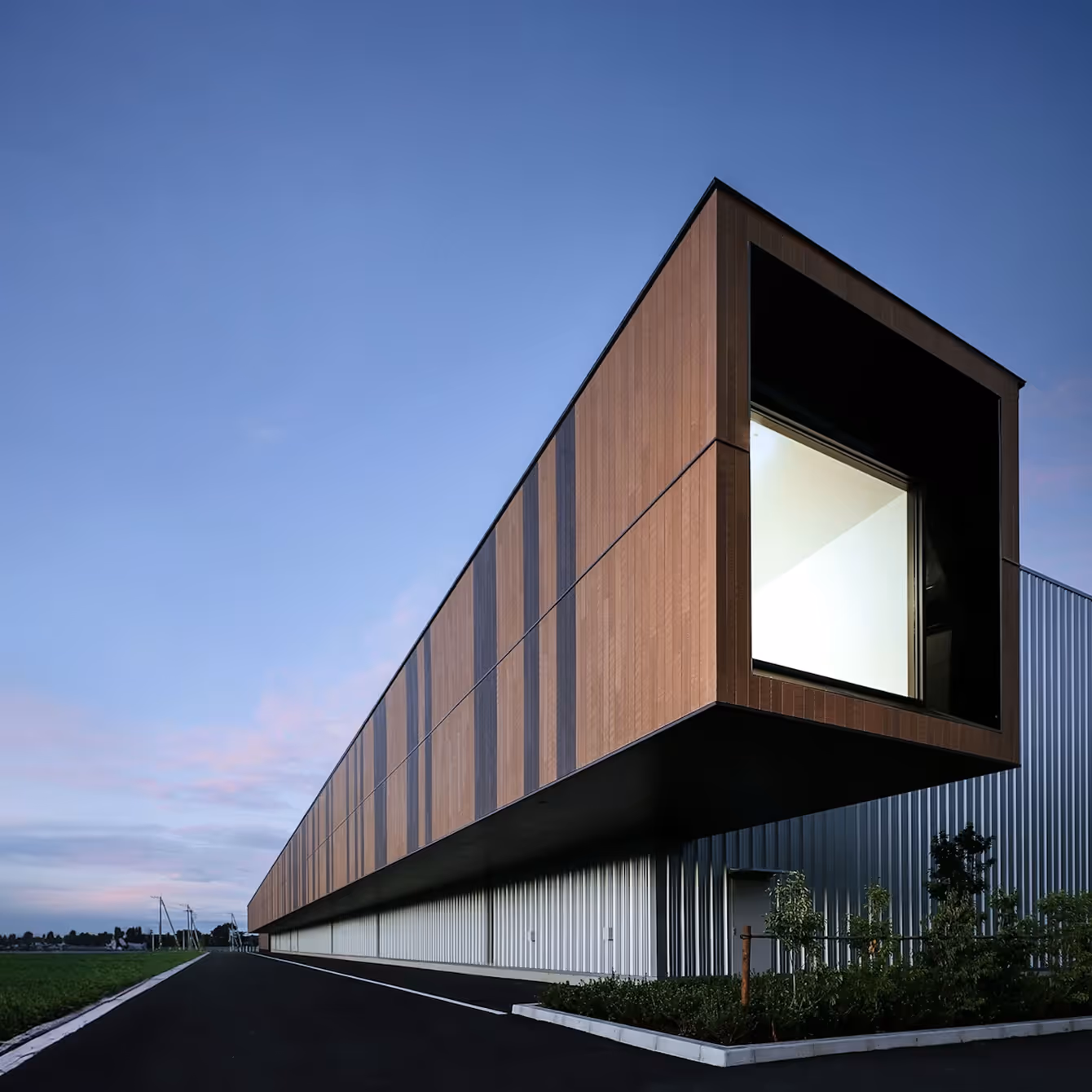 Modern building with an extended rectangular wooden facade and large window at dusk.