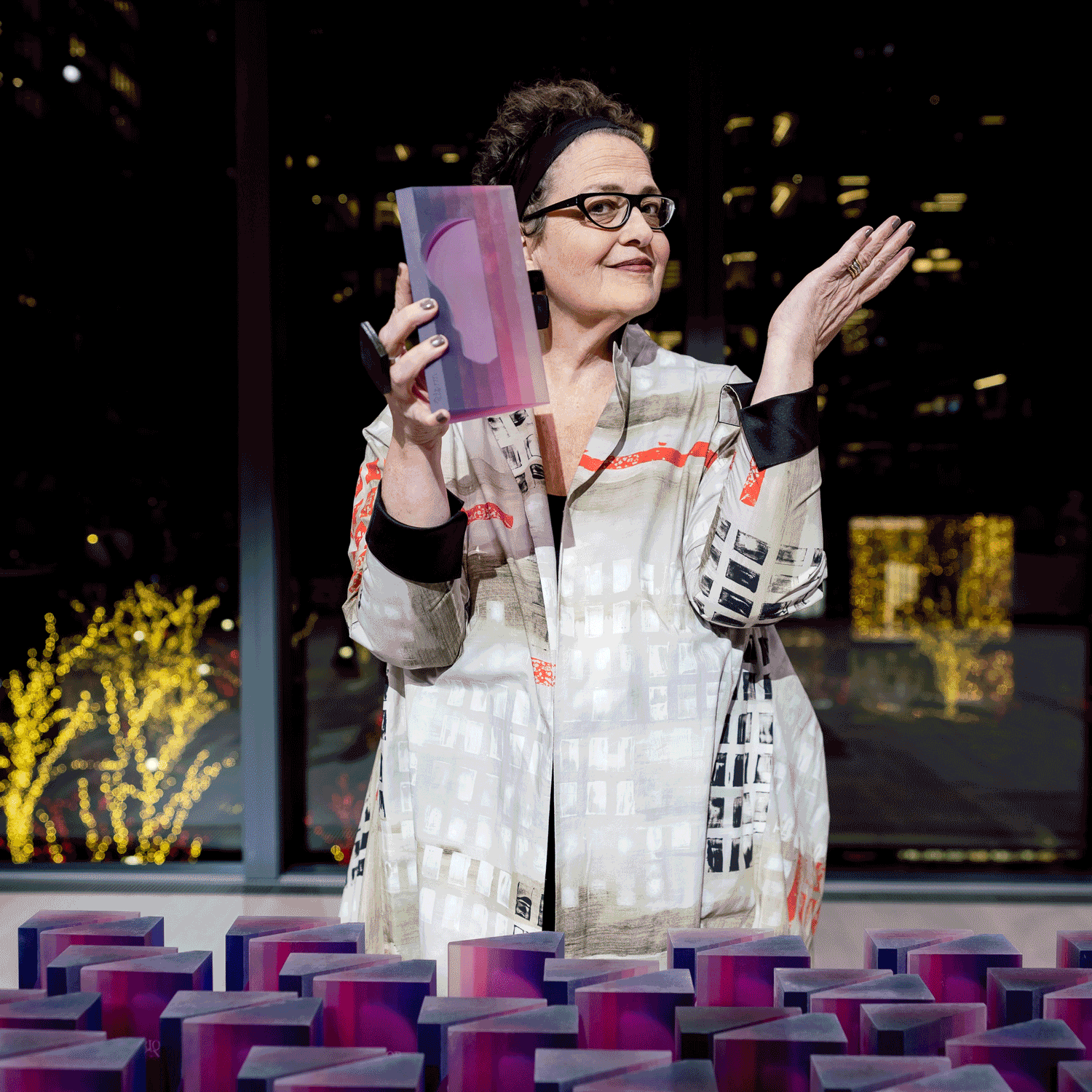 Woman wearing glasses and a patterned jacket holds a purple and pink rectangular award, standing behind a table filled with similar awards, with city lights in the background.