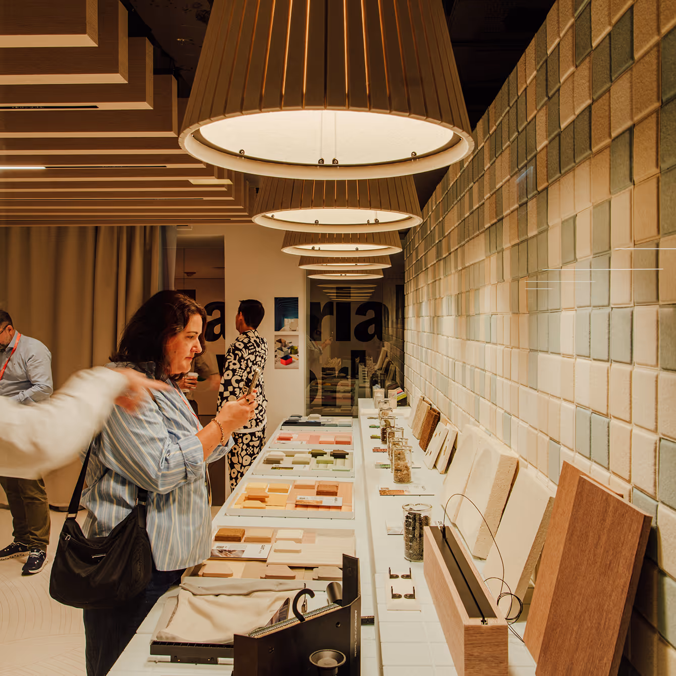 Woman observing design samples and materials displayed on a long table under large pendant lights in a modern showroom.