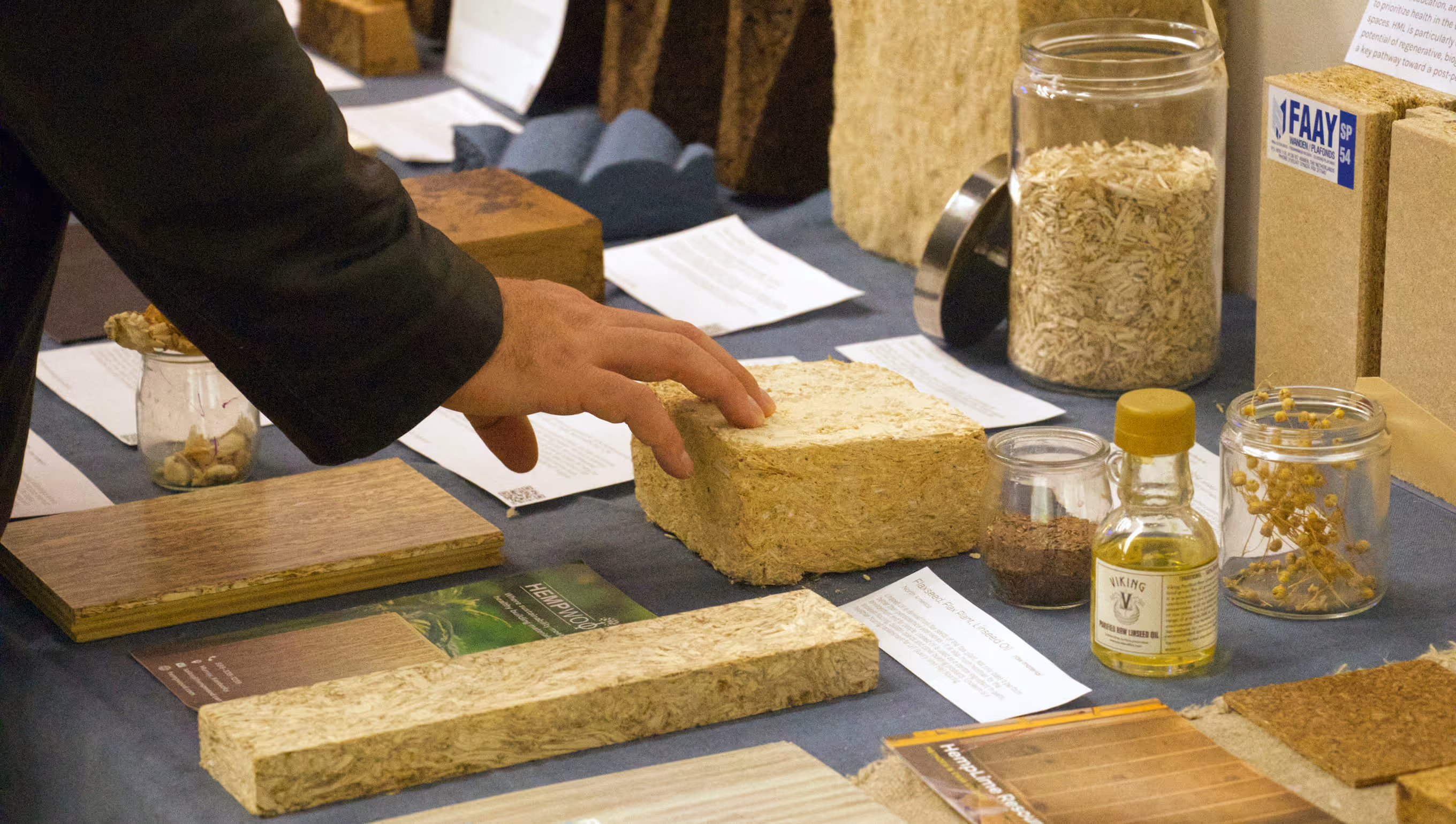 Hand touching a square block of compressed natural fibers on a table with various samples of wood, oil, and plant materials.