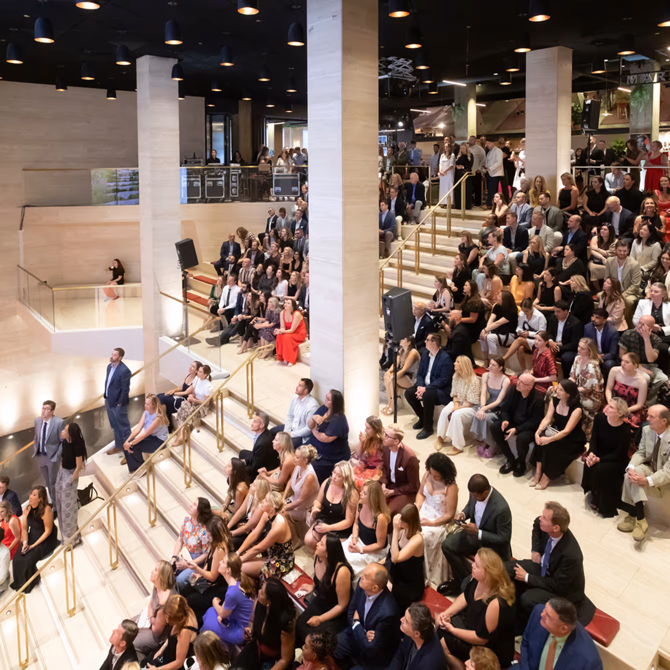 Large crowd of well-dressed people seated on tiered steps in a modern indoor venue, attentively watching an event.