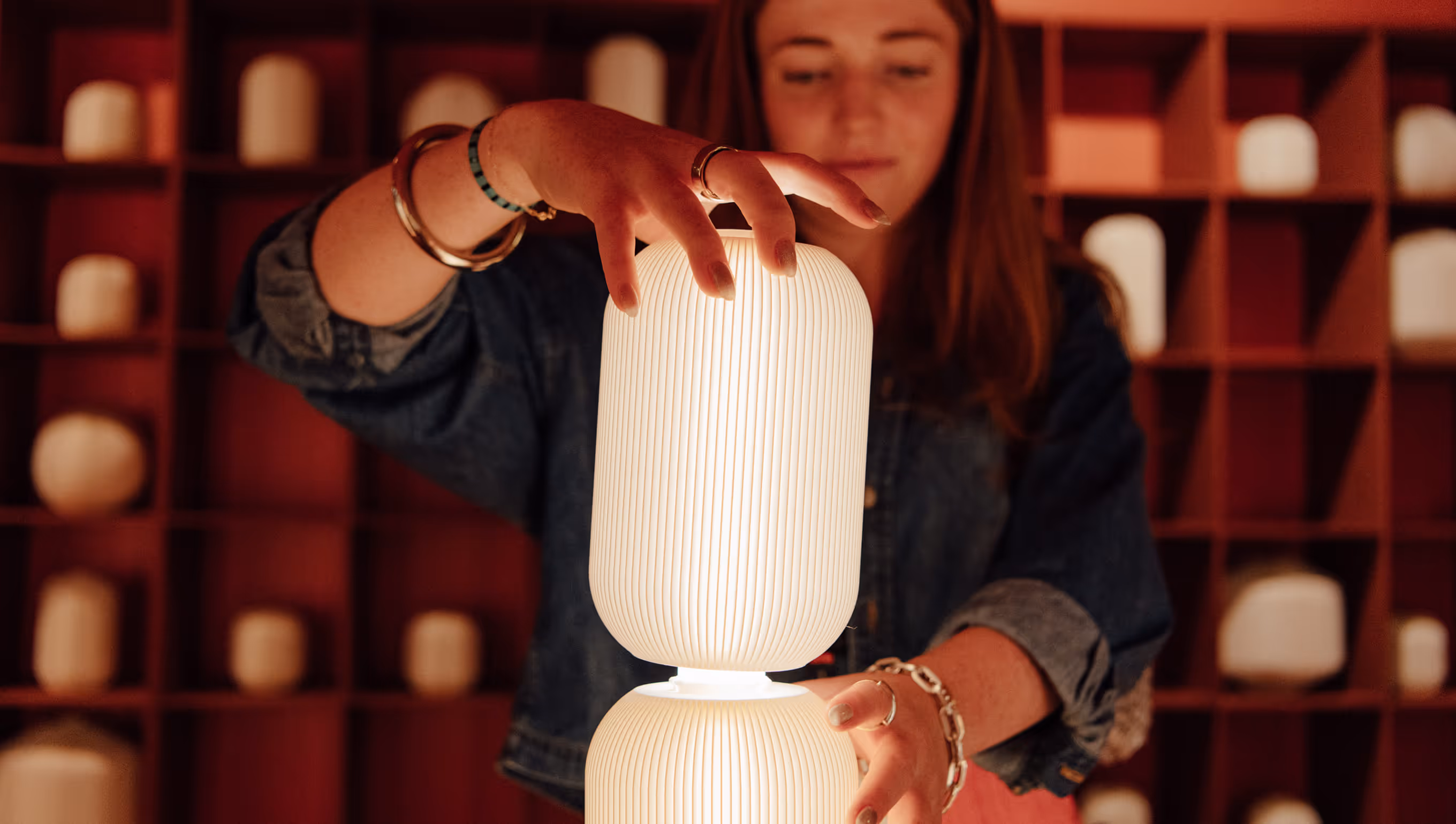 Woman adjusting a modern, white, ribbed table lamp with soft warm light in front of a wooden shelf with similar lamps.