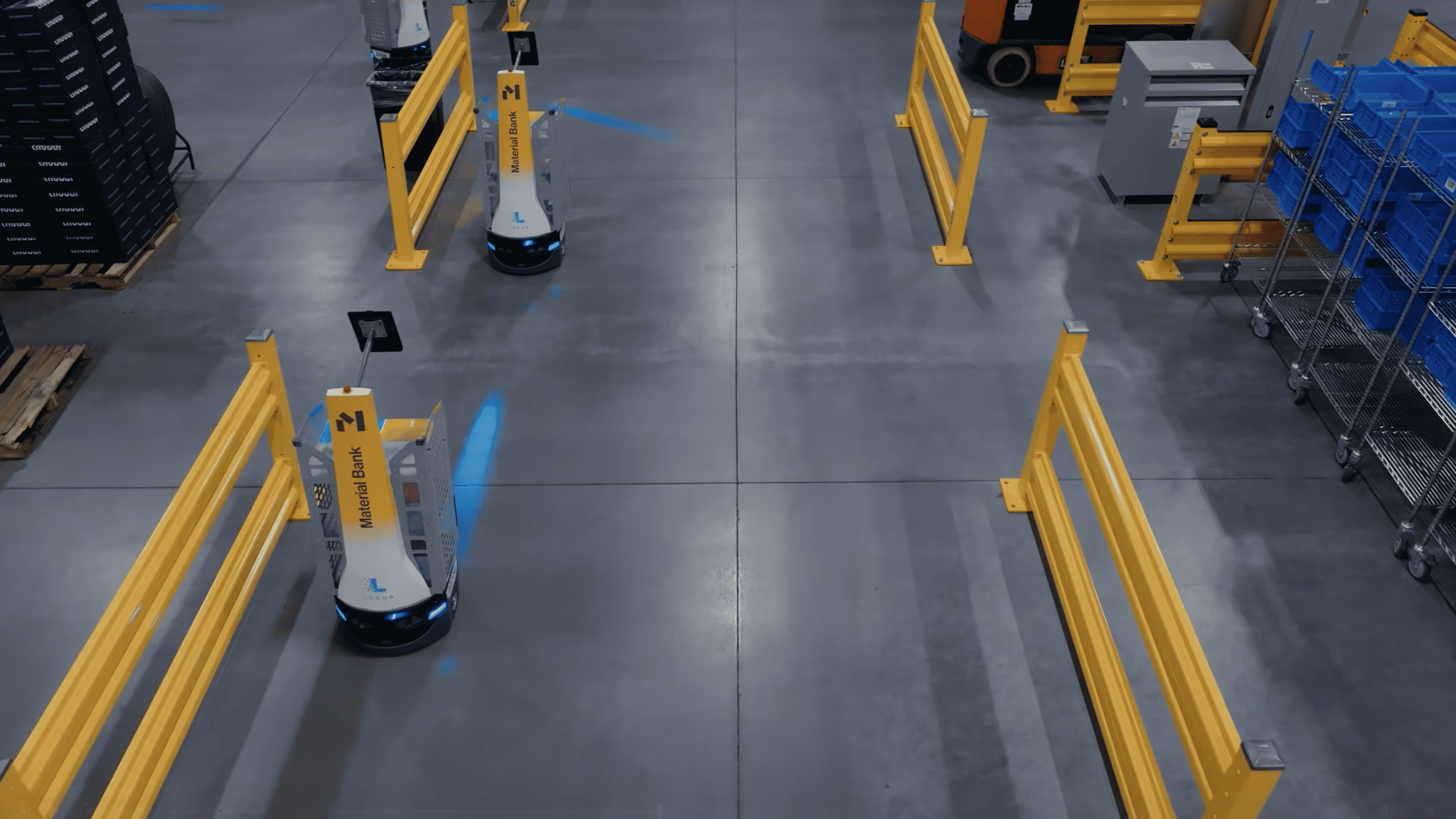 Two autonomous mobile robots labeled 'Material Bank' navigating in an industrial warehouse with yellow safety barriers.