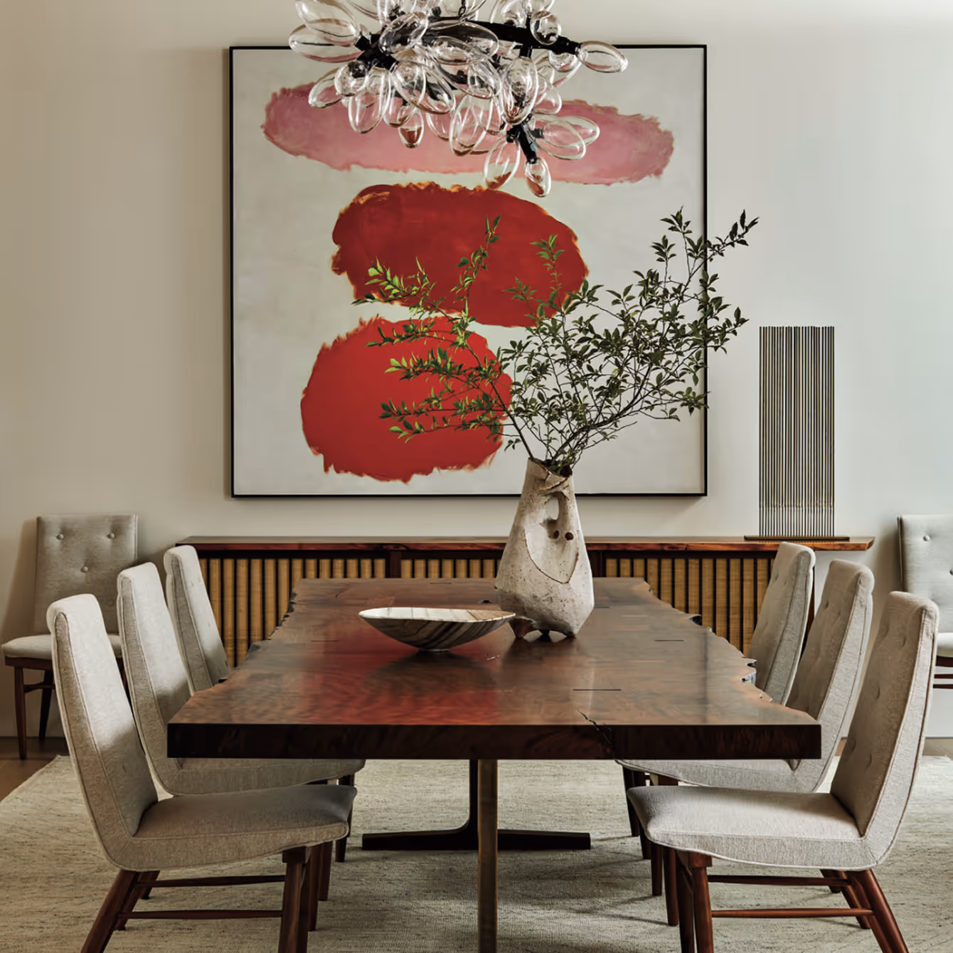 Modern dining room with a wooden table, six beige upholstered chairs, a sculptural vase with greenery, abstract red artwork, and a glass chandelier.