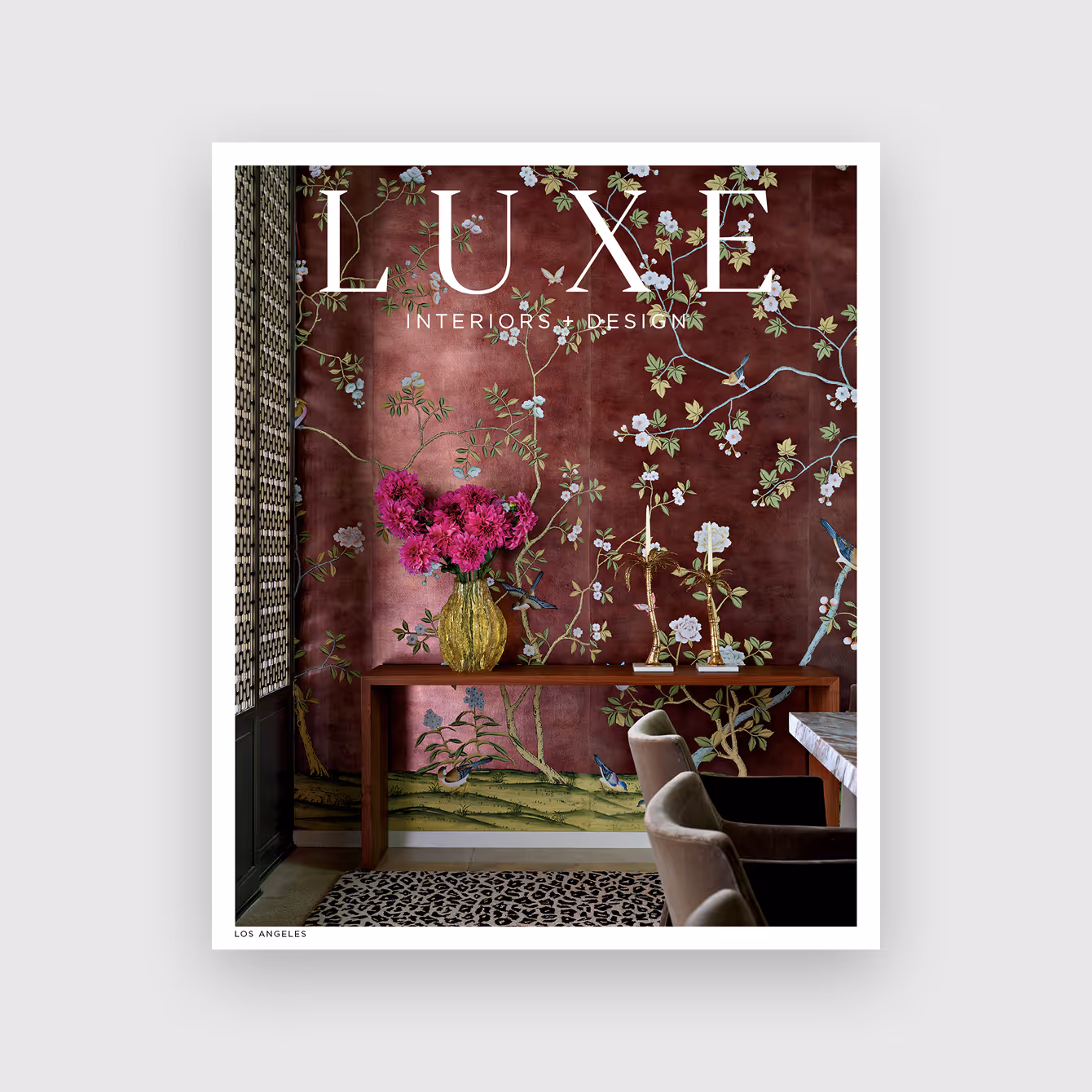 Luxe Interiors + Design magazine cover featuring a floral wallpapered room with a wooden console table holding a yellow vase with magenta flowers and gold candlesticks, beside velvet dining chairs and a leopard print rug.