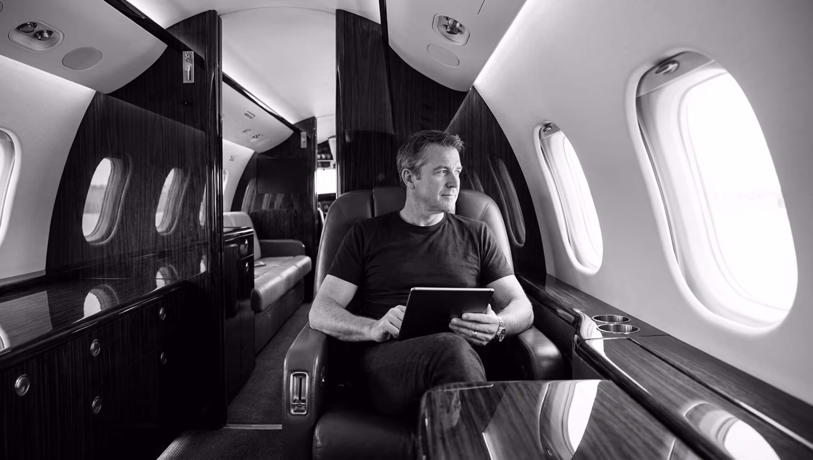 Man seated in a leather chair inside a private jet, holding a tablet and looking out the window.