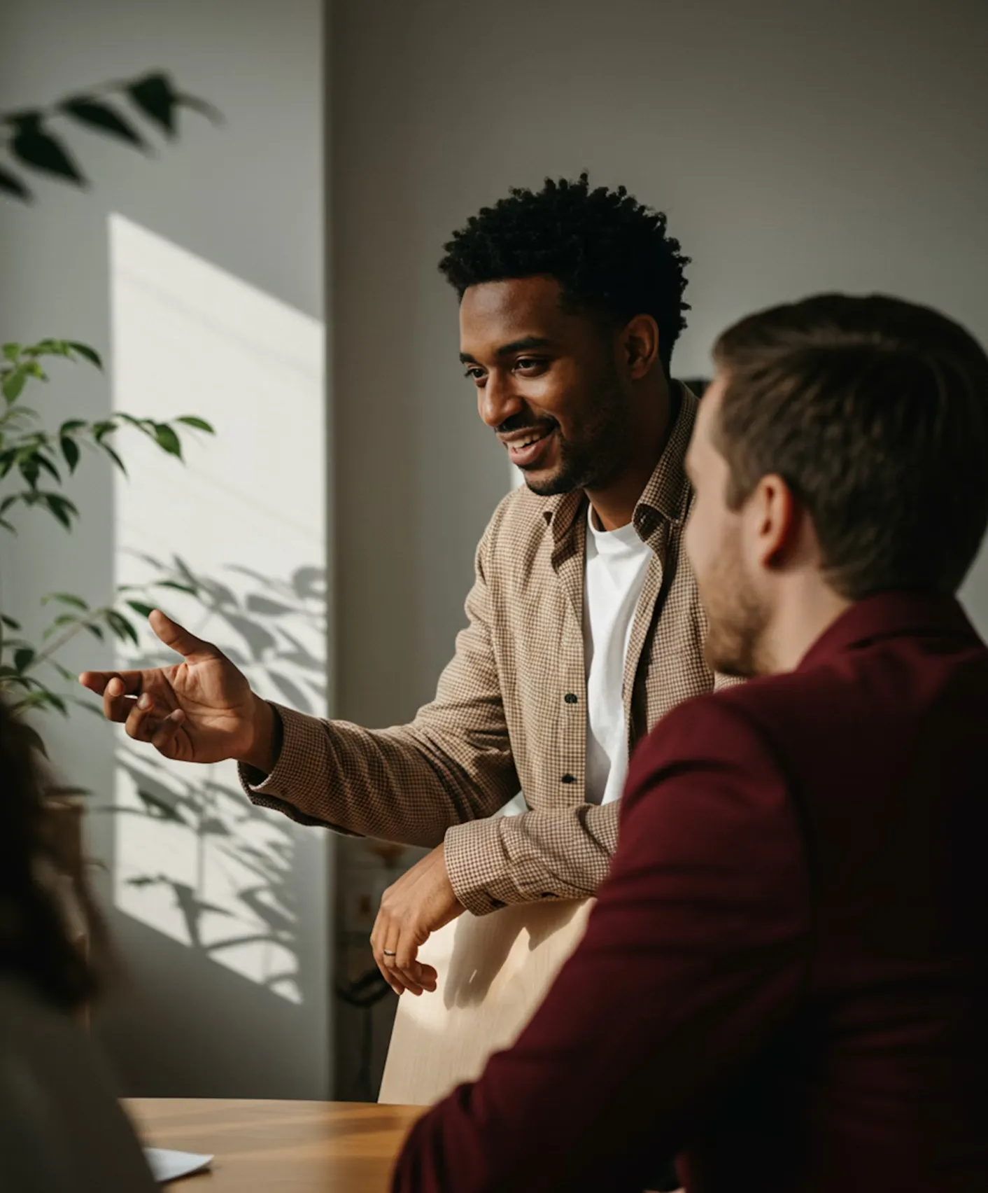 Man with curly hair wearing a checked shirt smiling and gesturing while talking to two others in a sunlit room.