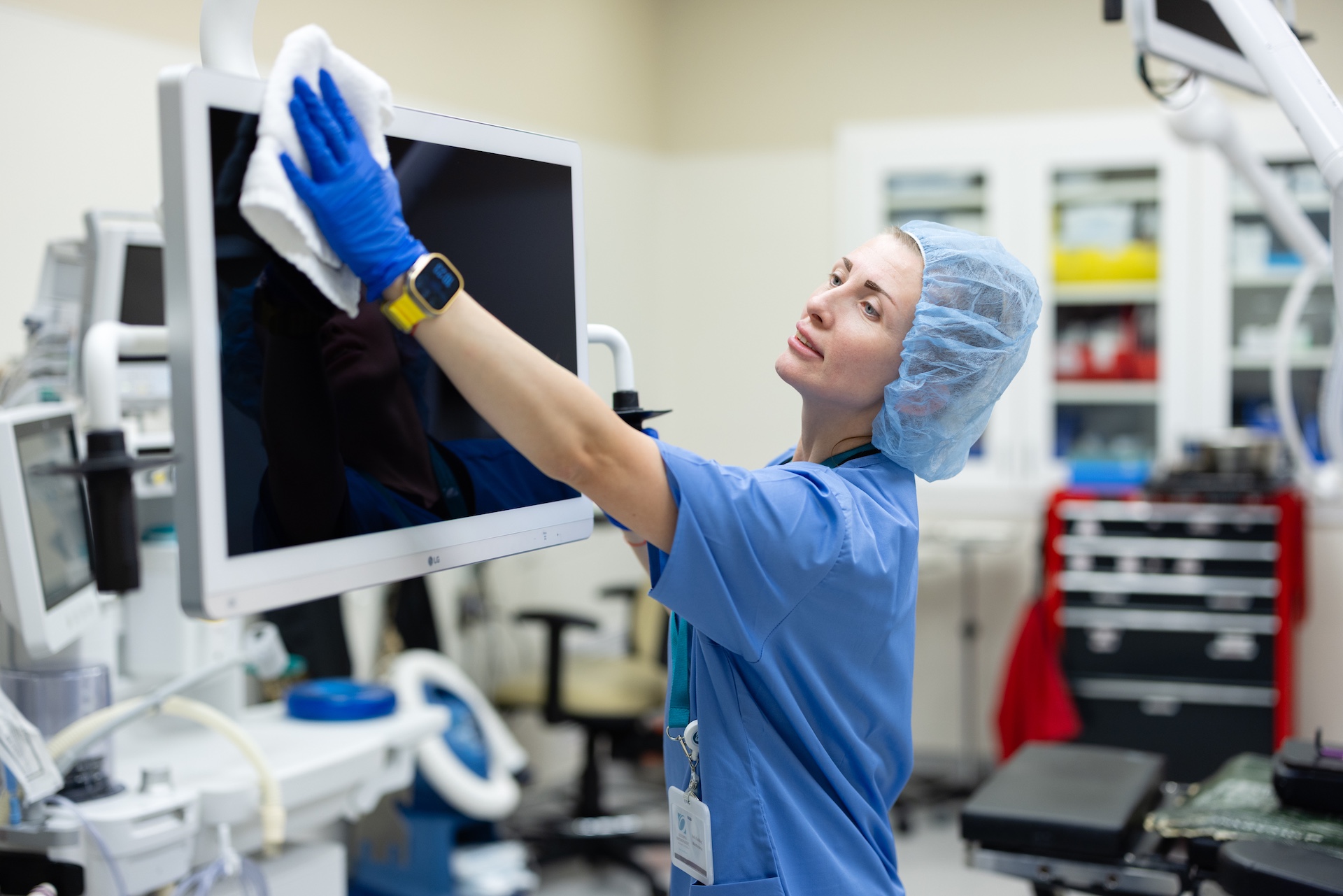 Healthcare worker in scrubs and a hair cover cleaning a medical monitor in a hospital room.