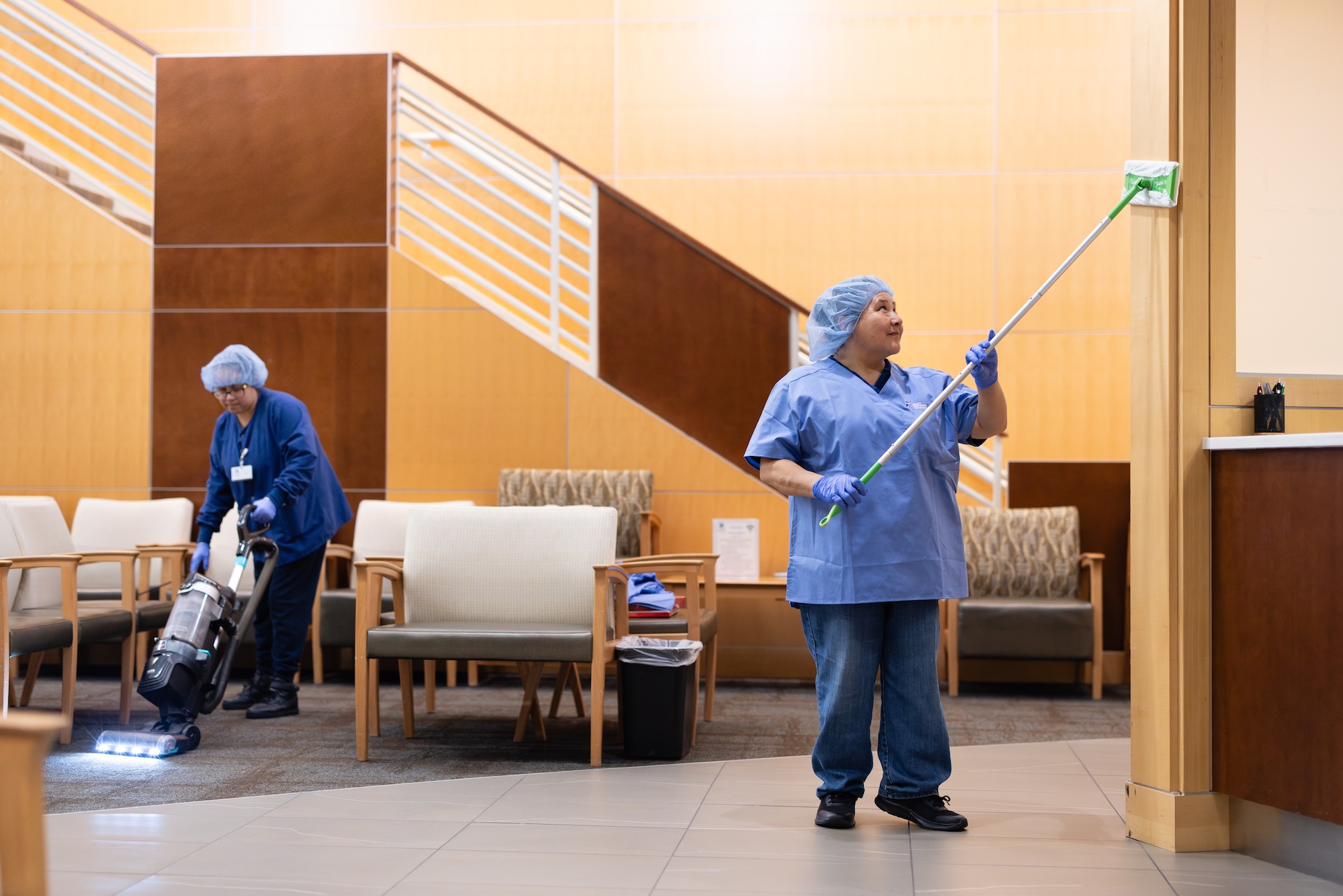 Two janitors cleaning a hallway; one is vacuuming the carpet while the other dusts a wooden wall with a long-handled duster.