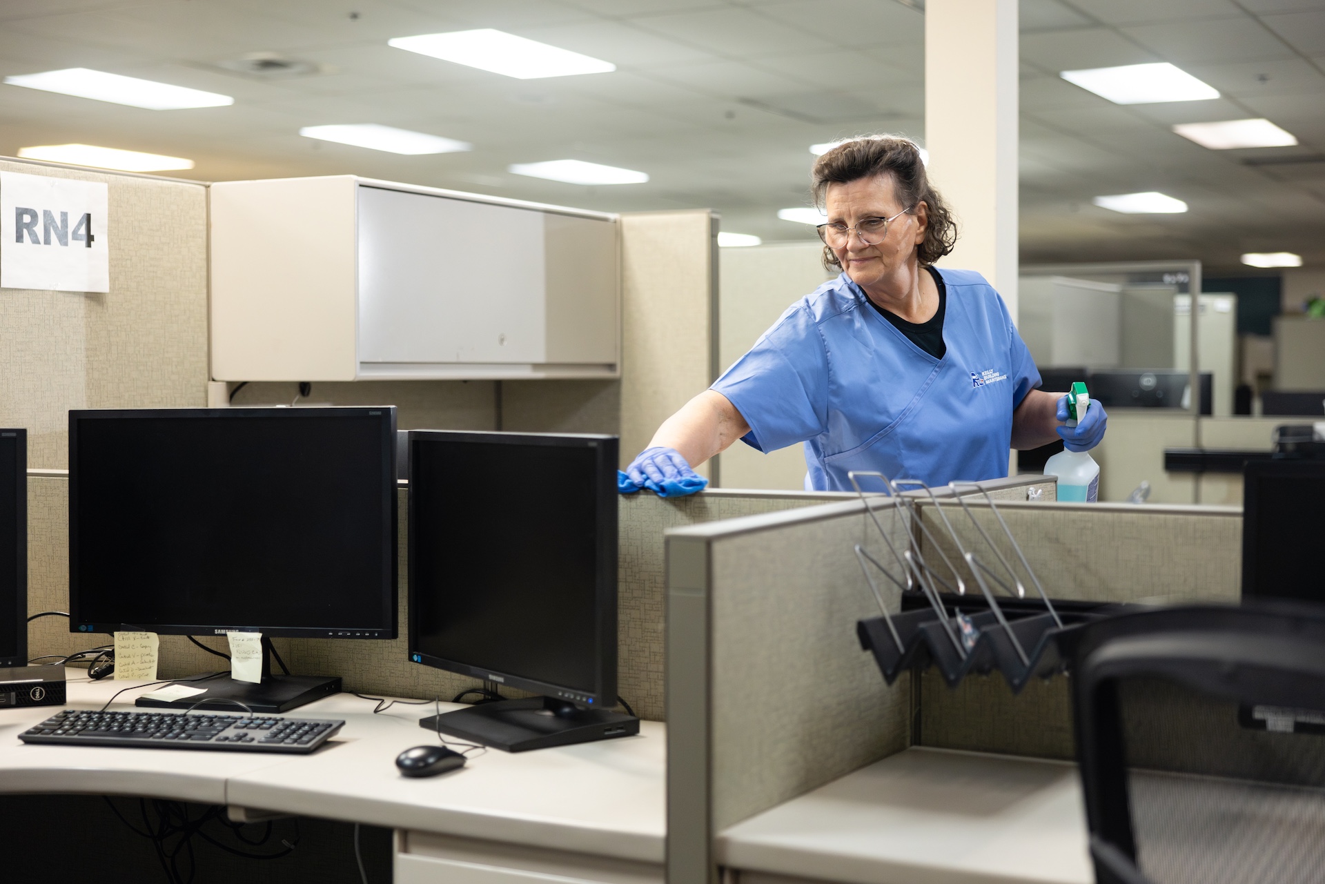 Woman in blue scrubs and gloves cleaning an office cubicle desk with spray bottle and cloth.