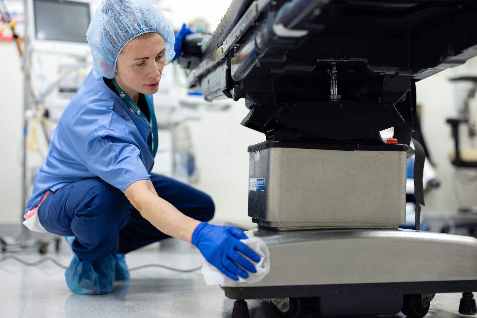Healthcare worker in blue scrubs and gloves cleaning under a hospital bed in a medical facility.
