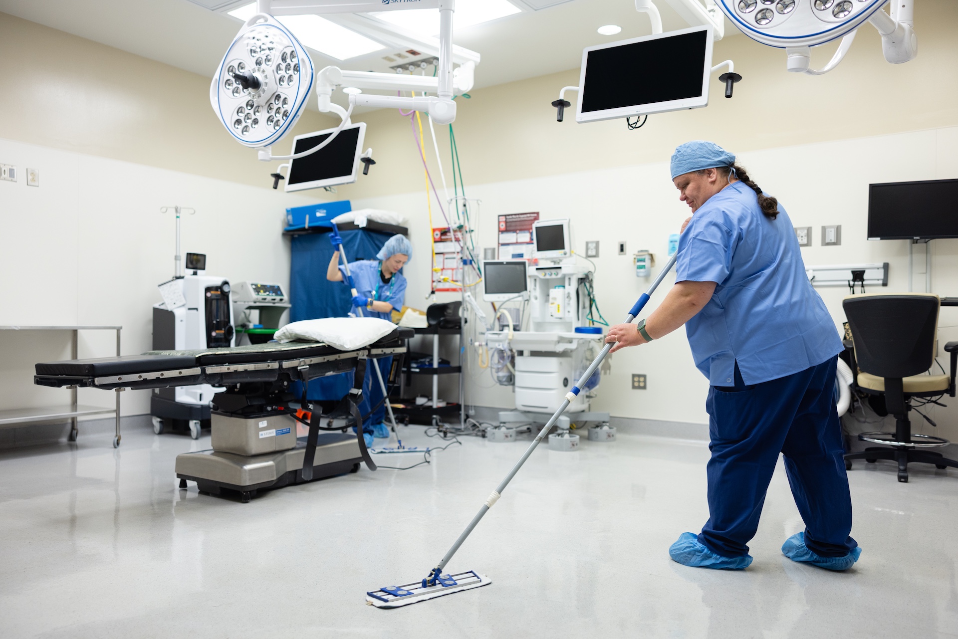 Two healthcare workers in blue scrubs cleaning the floor of a hospital operating room with mops.