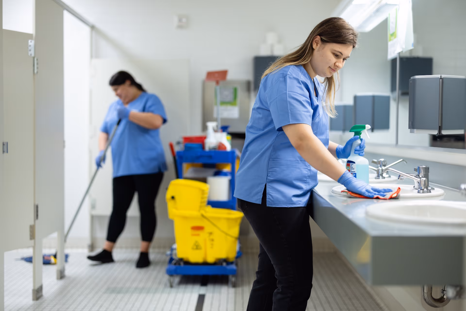 Two janitors in blue uniforms cleaning a public restroom with one wiping the sink and the other mopping the floor near cleaning supplies.
