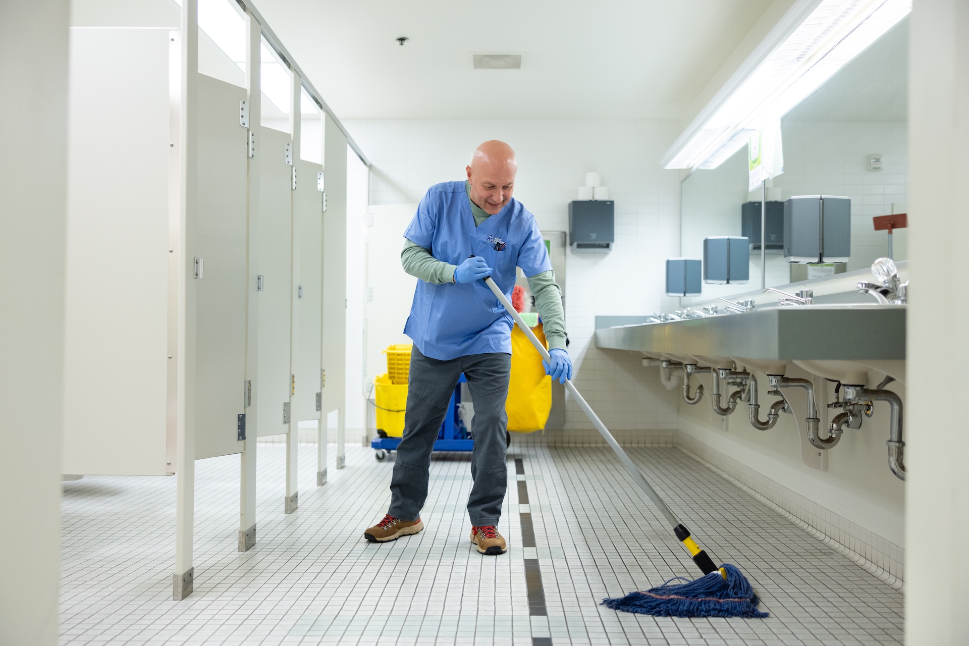 Janitor mopping the tiled floor of a clean public restroom with sinks and stalls.