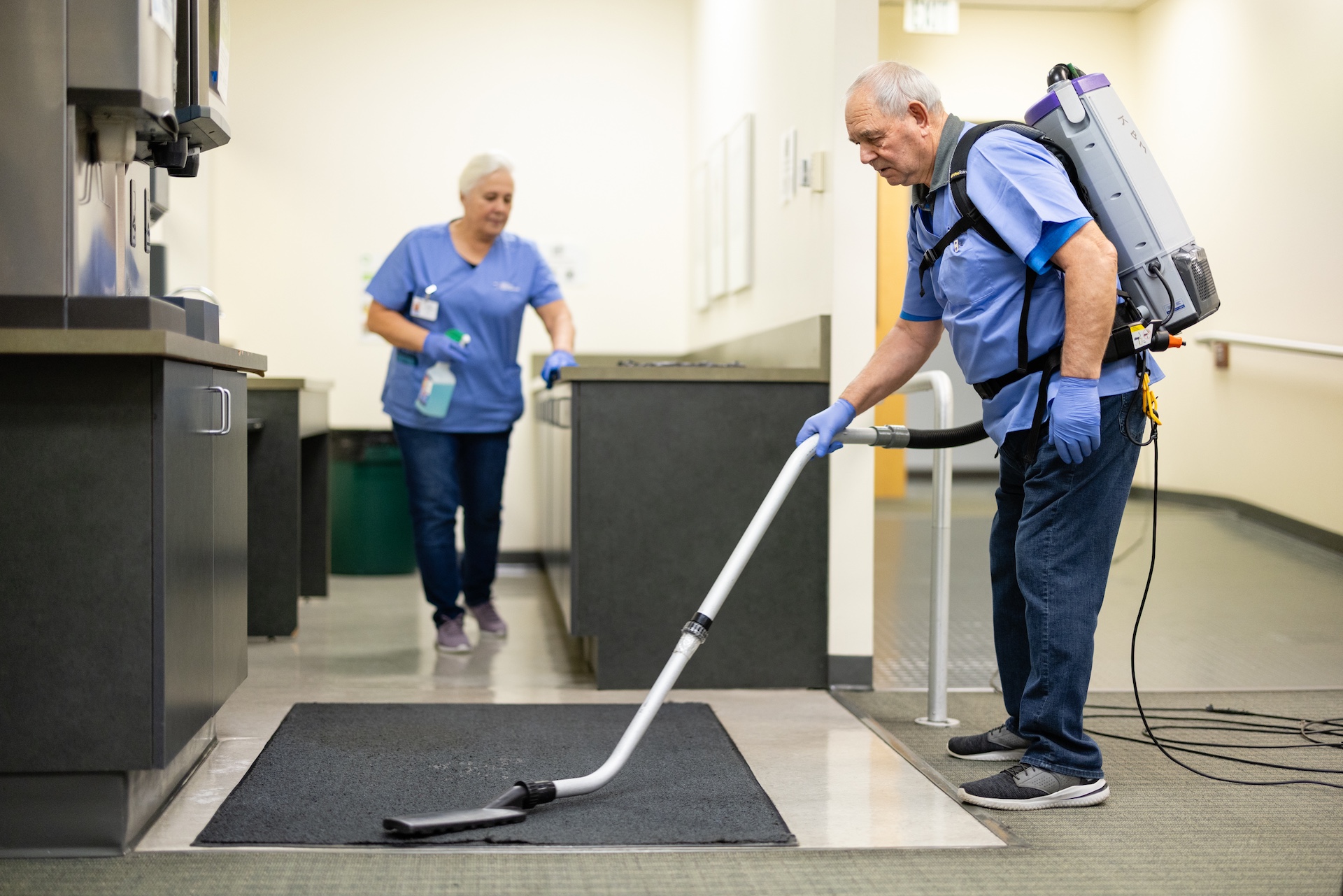 Two janitors wearing blue uniforms cleaning a room, one vacuuming a floor mat and the other holding a spray bottle.