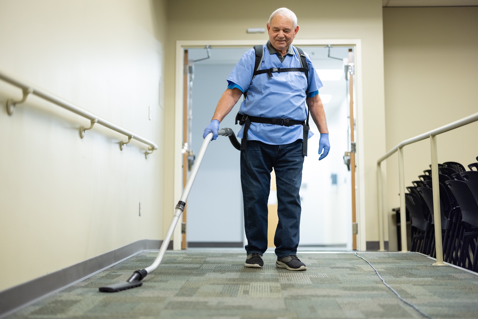 Older man in blue shirt and gloves vacuuming carpeted floor in a hallway with handrails.