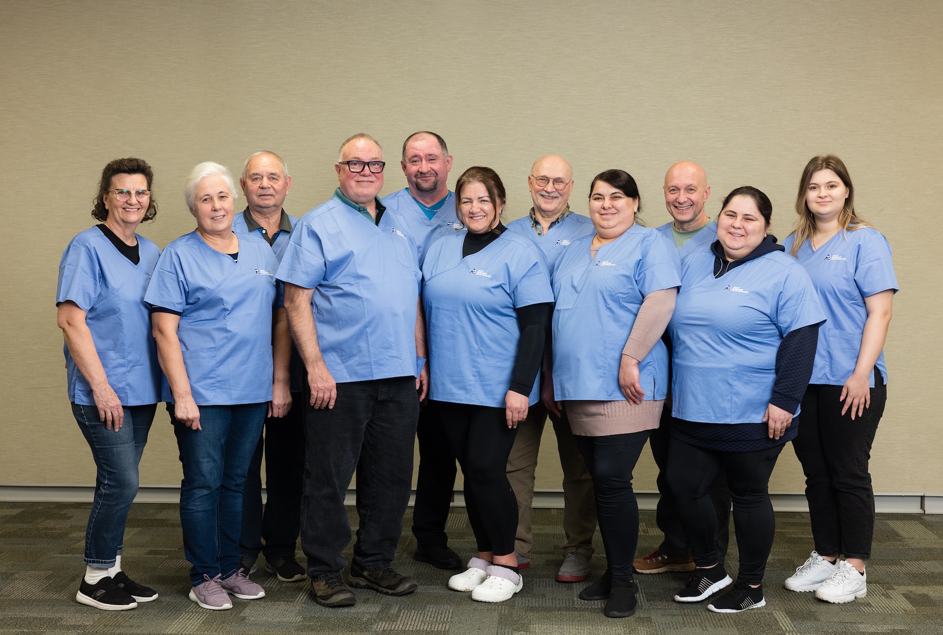 Group of ten adults wearing light blue medical scrubs standing indoors against a beige wall, smiling at the camera.