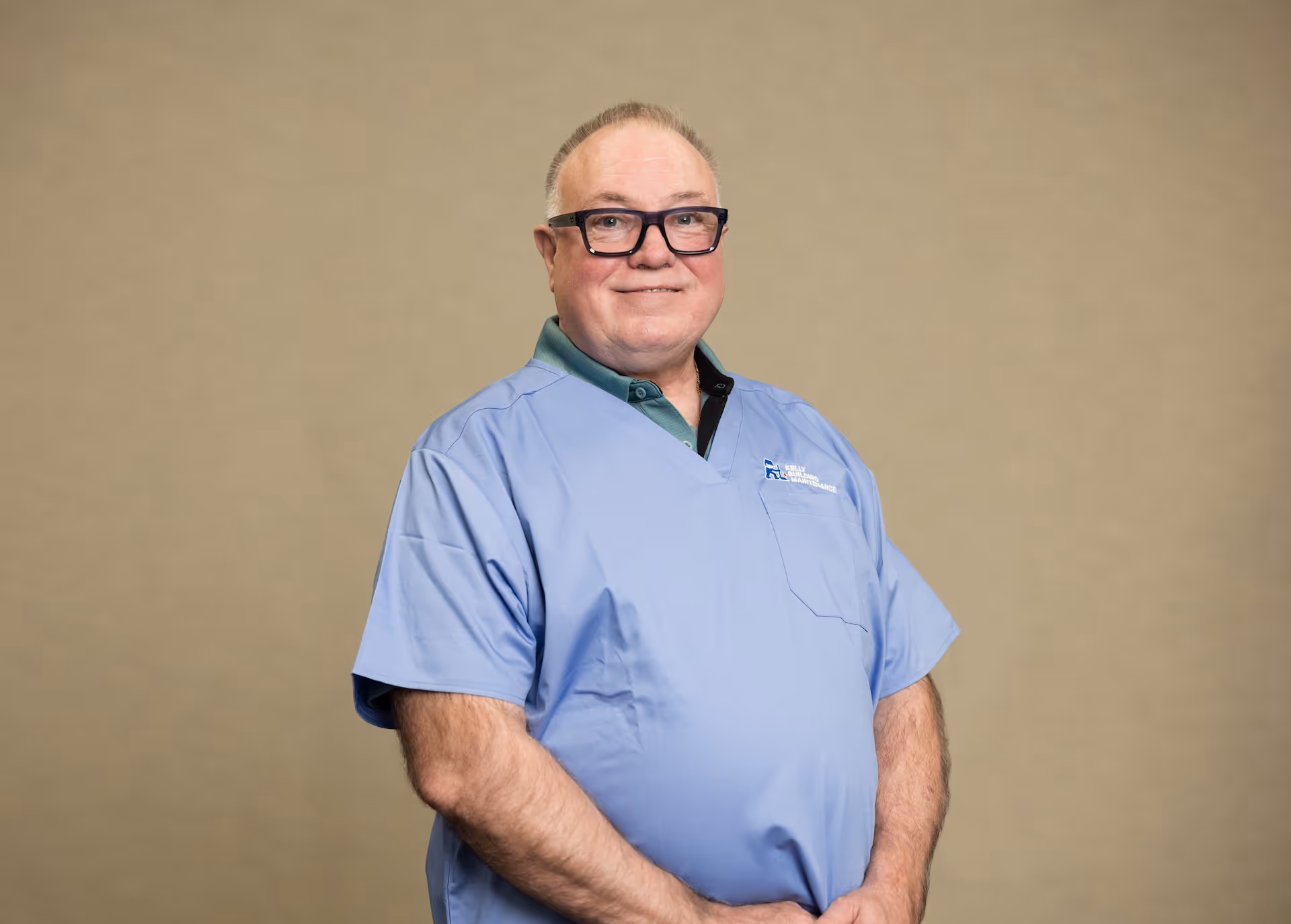 Smiling man in blue medical scrubs and glasses standing against a plain beige background.