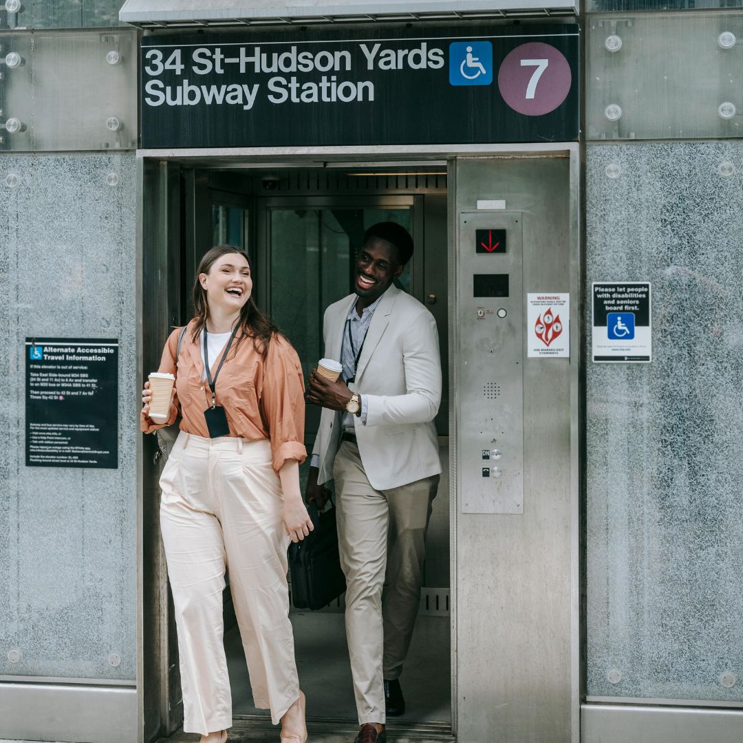 Two smiling people holding coffee cups exiting the 34 St-Hudson Yards Subway Station elevator for the 7 line.