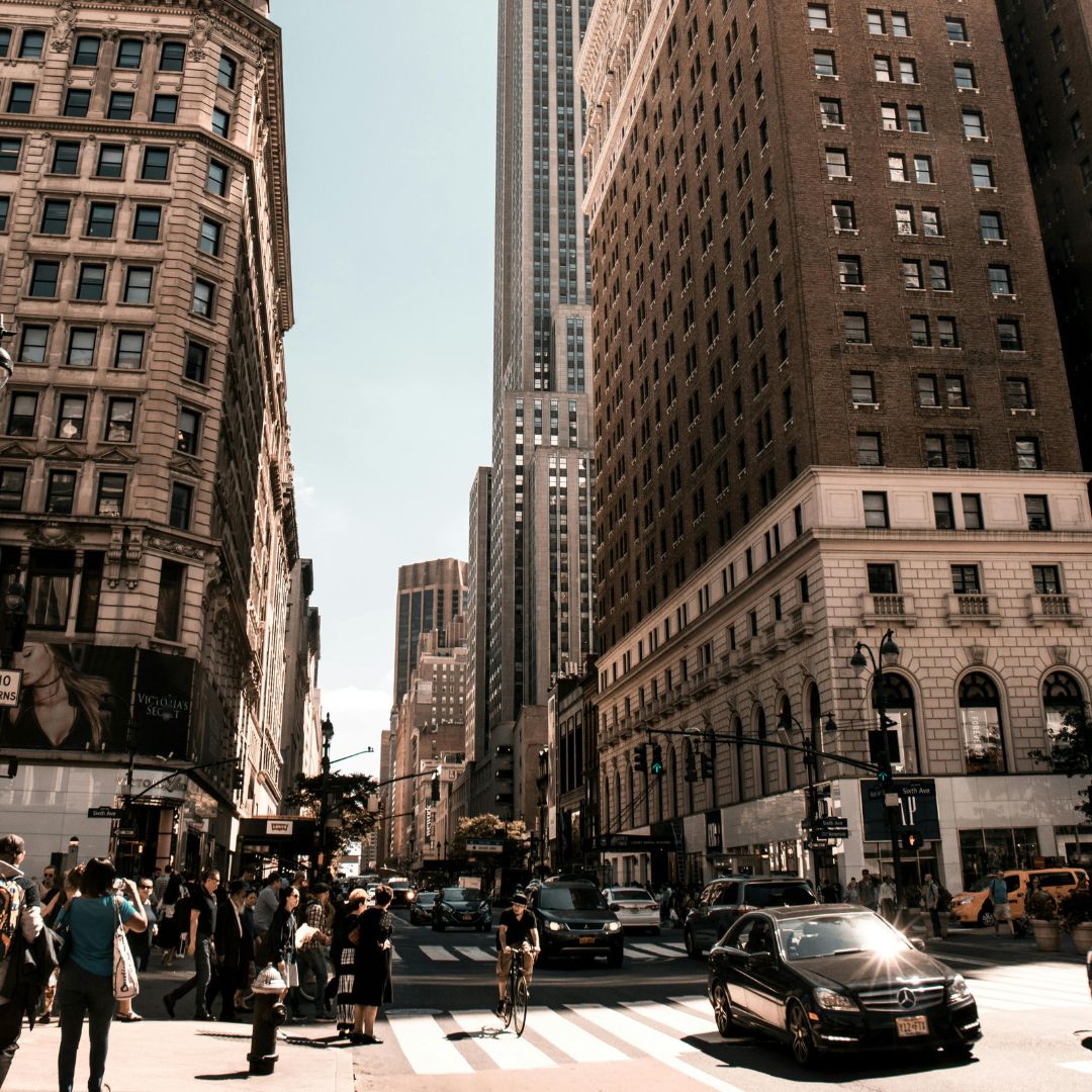 Busy city street with tall buildings, pedestrians waiting to cross, and vehicles including a cyclist and a black car.