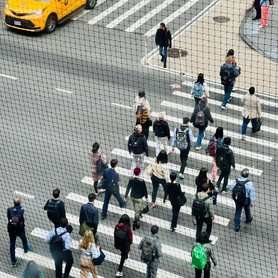 Overhead view of diverse pedestrians crossing a city street at a crosswalk with a yellow taxi nearby, seen through a safety net.