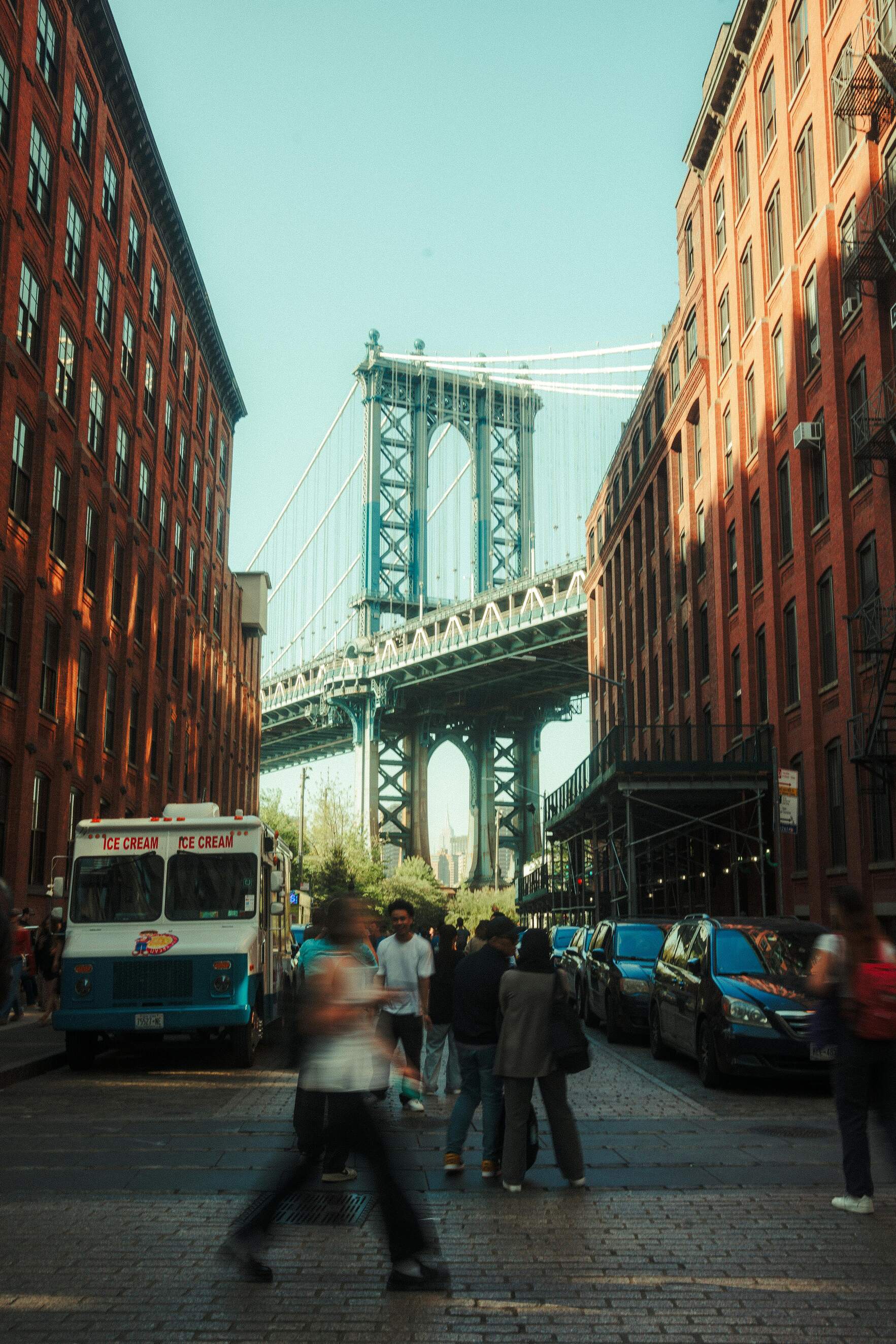 View of the Manhattan Bridge framed by red brick buildings with people walking and an ice cream truck parked on the street.