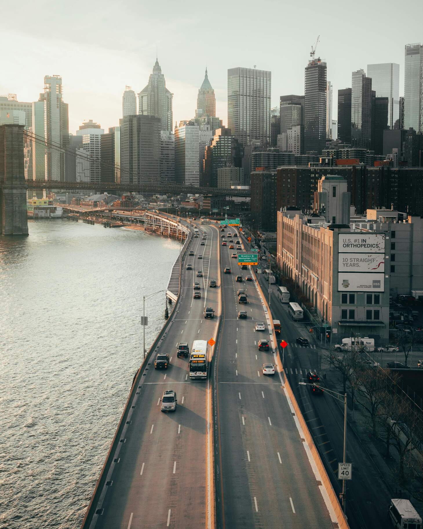 Elevated highway with vehicles leading into a dense city skyline by a river during sunset.