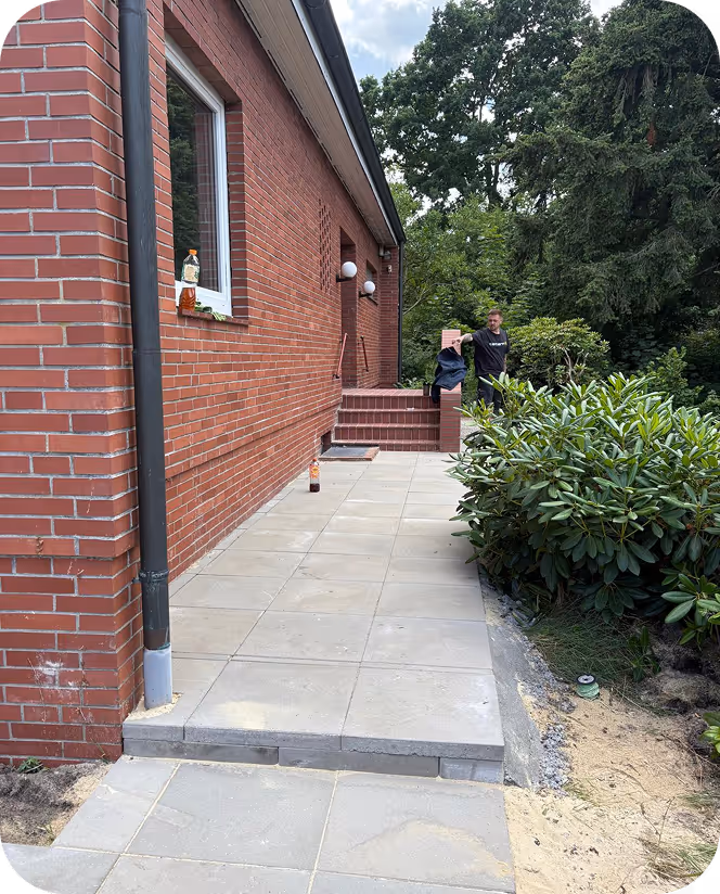 Ein Mann steht auf einer neu gepflasterten Terrasse neben einem roten Backsteinhaus mit Pflanzen rechts und Bäumen im Hintergrund.