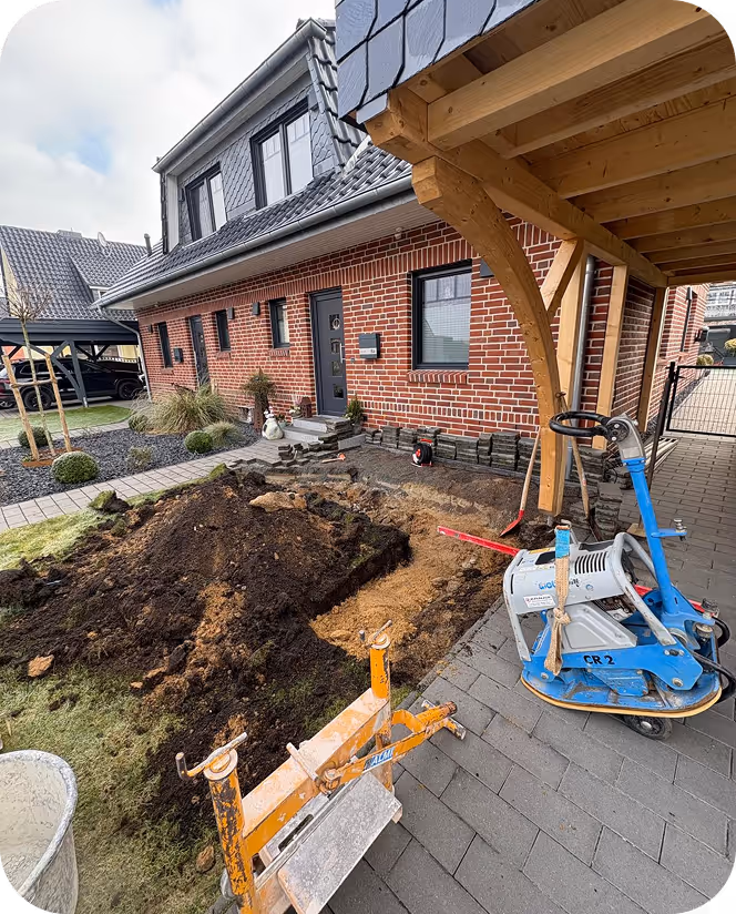 Baustelle vor einem modernen roten Backsteinhaus mit einem aufgebaggerten Bereich und Baumaschinen unter einem Holzcarport.