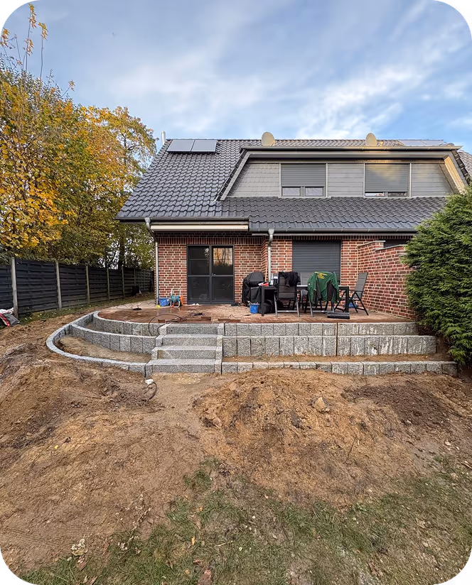 Ein zweistöckiges Backsteinhaus mit grauem Ziegeldach, einer Terrasse mit Tisch und Stühlen und einer neu gebauten Stützmauer aus Steinblöcken im Vordergrund.