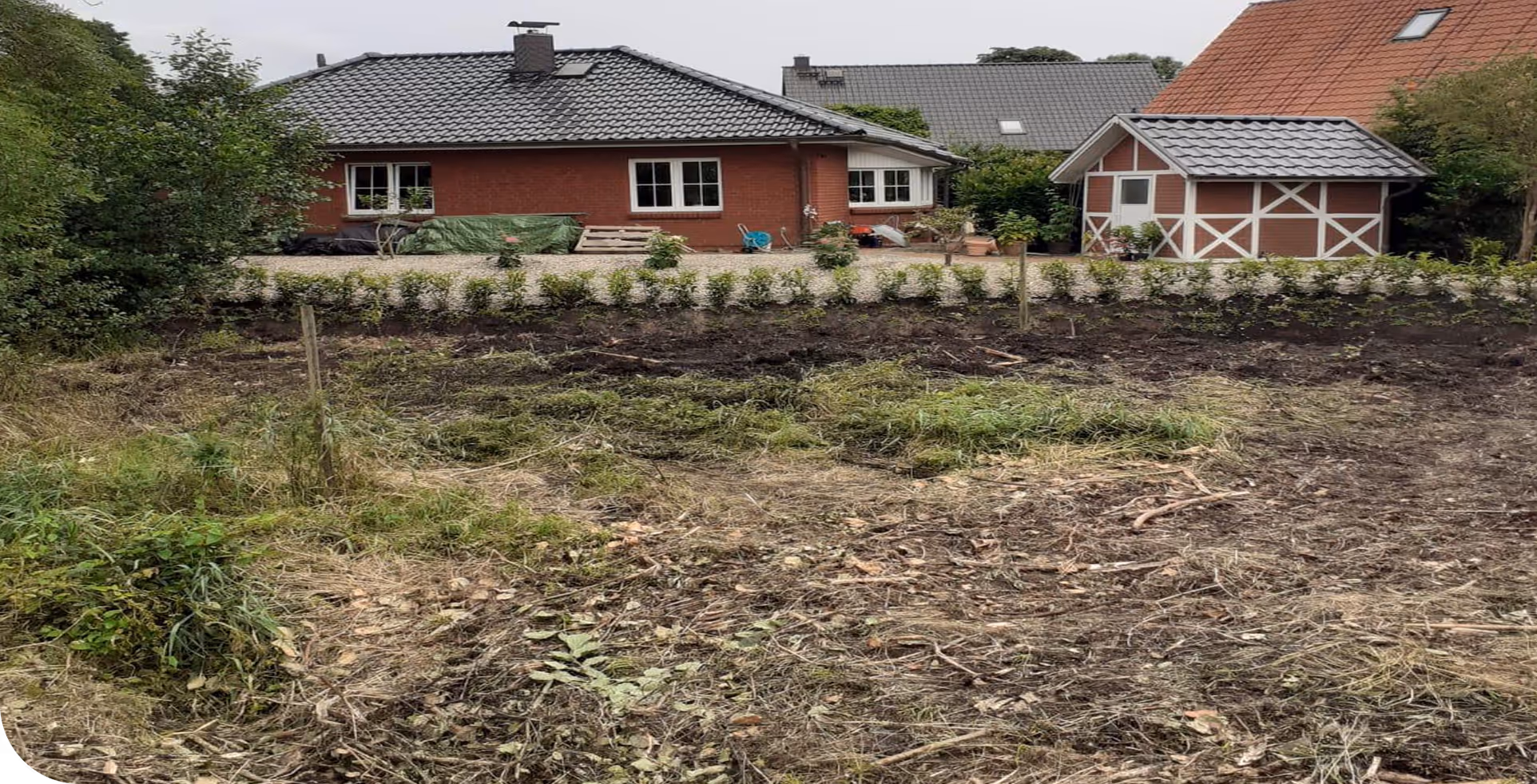Unbebautes, teilweise brachliegendes Grundstück mit unordentlichem Gras und Erde vor zwei Einfamilienhäusern mit Ziegeldächern.