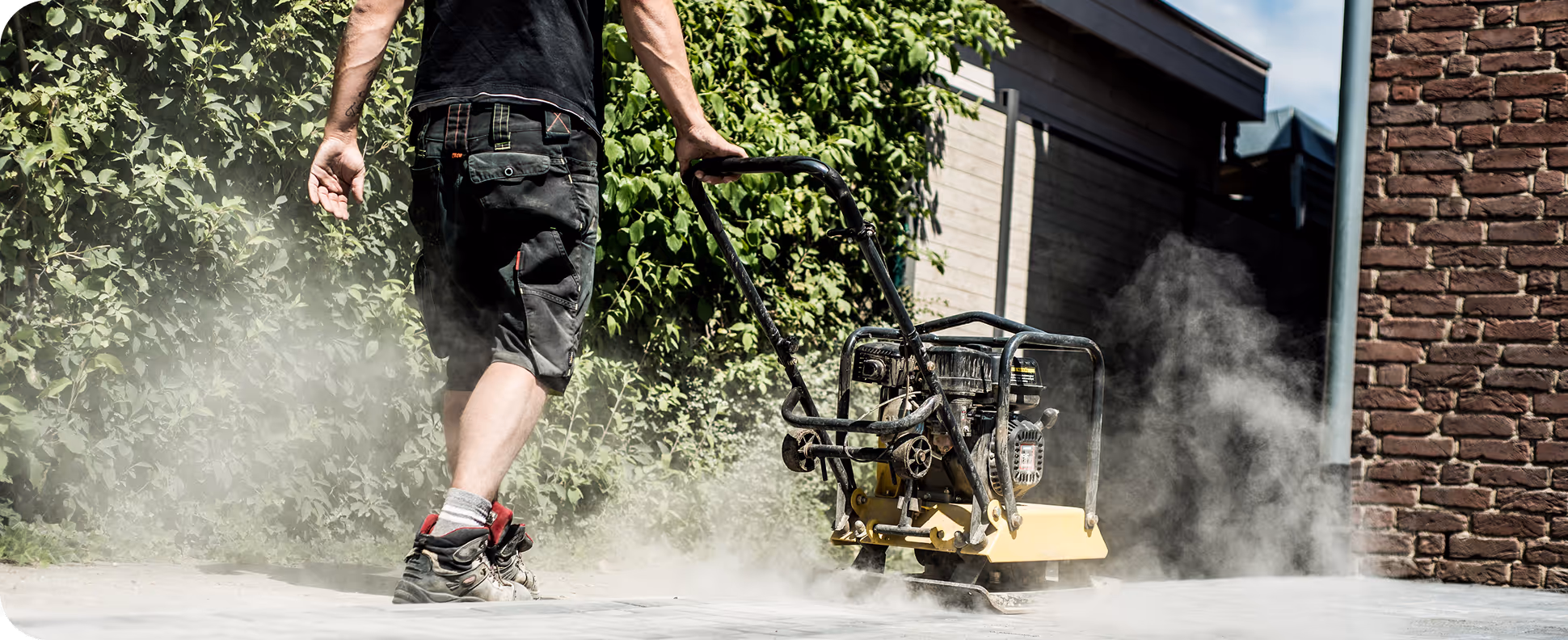 Person bedient eine Bodenverdichtungsmaschine auf einem staubigen Gehweg neben einer grünen Hecke und einem Backsteingebäude.