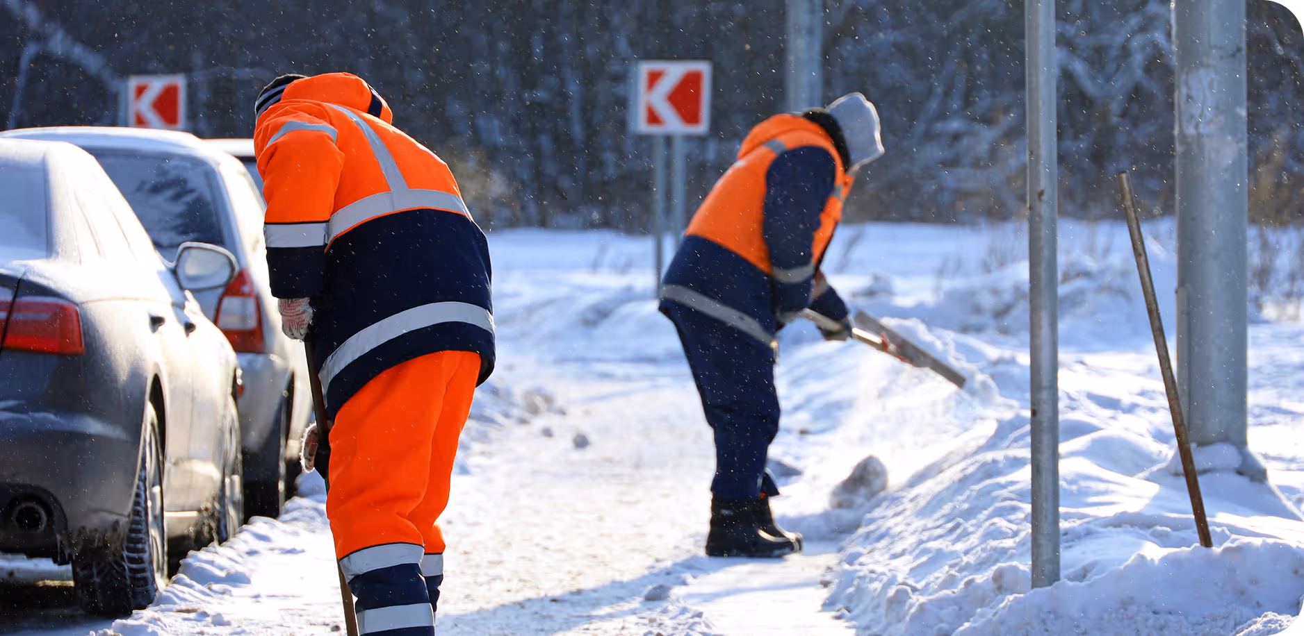Zwei Personen in orange-schwarzer Winterarbeitskleidung räumen Schnee von einem Gehweg neben parkenden Autos.