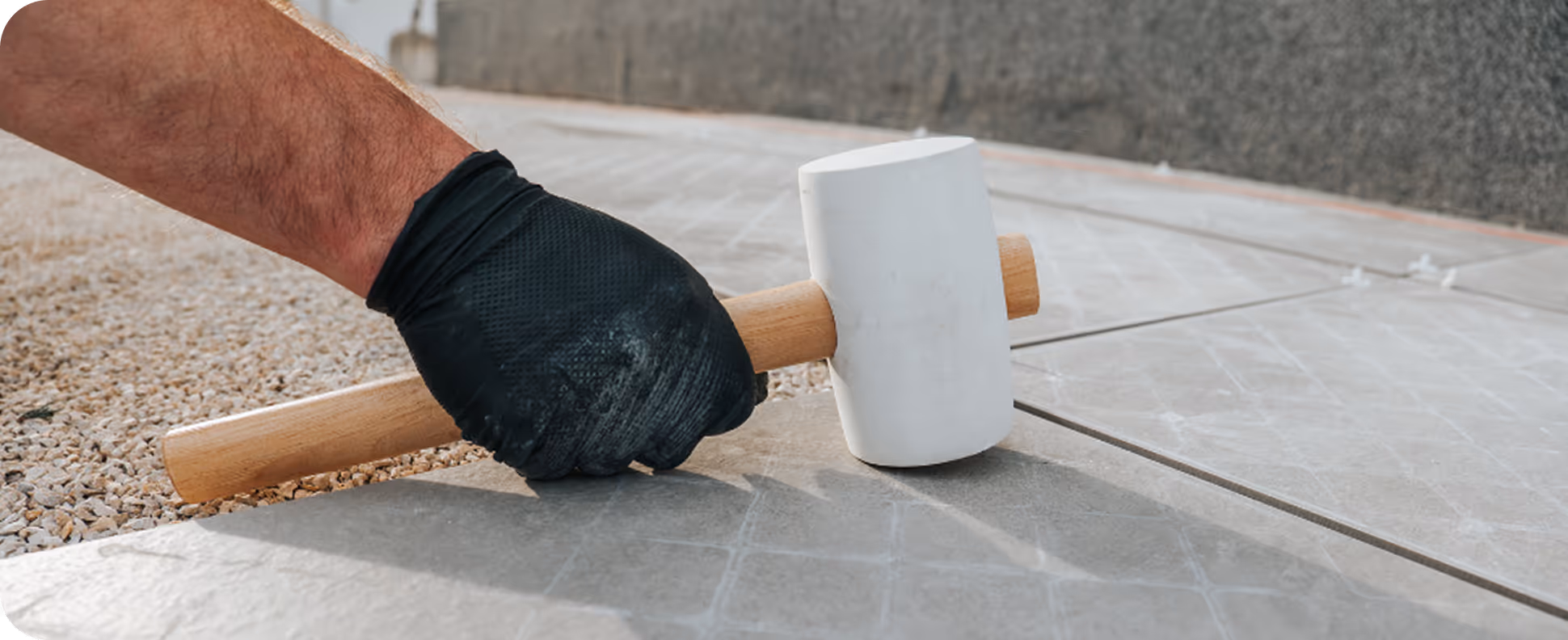 Hand in schwarzem Handschuh hält einen weißen Gummihammer über graue Bodenfliesen.