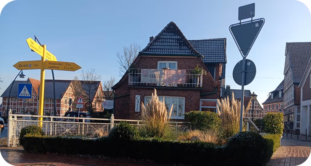 Straßenszene mit einem Fachwerkhaus, gelben Wegweisern, Verkehrszeichen und grüner Bepflanzung unter blauem Himmel.