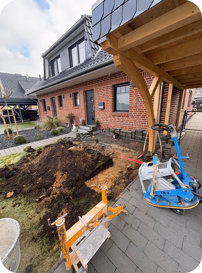 Baustelle vor einem roten Backsteinhaus mit einem aufgerissenen Erdreich und Baugeräten unter einer Holzkonstruktion.
