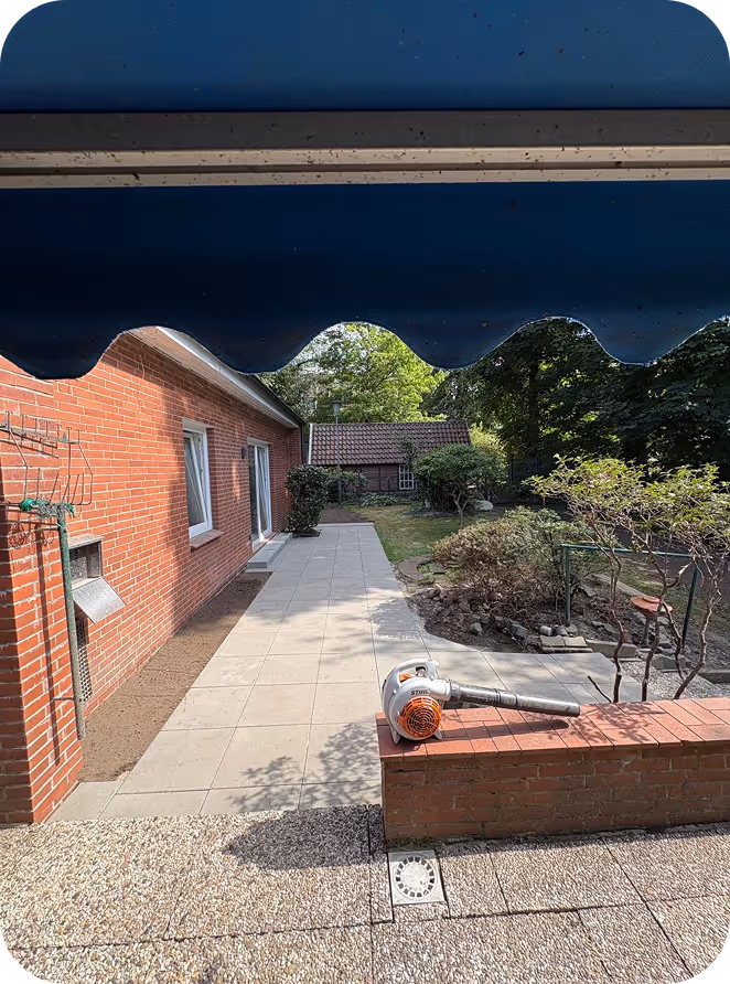 Freitagspatel mit blauer Markise, gepflasterter Terrasse neben rotem Backsteinhaus und Garten mit Büschen im Hintergrund.