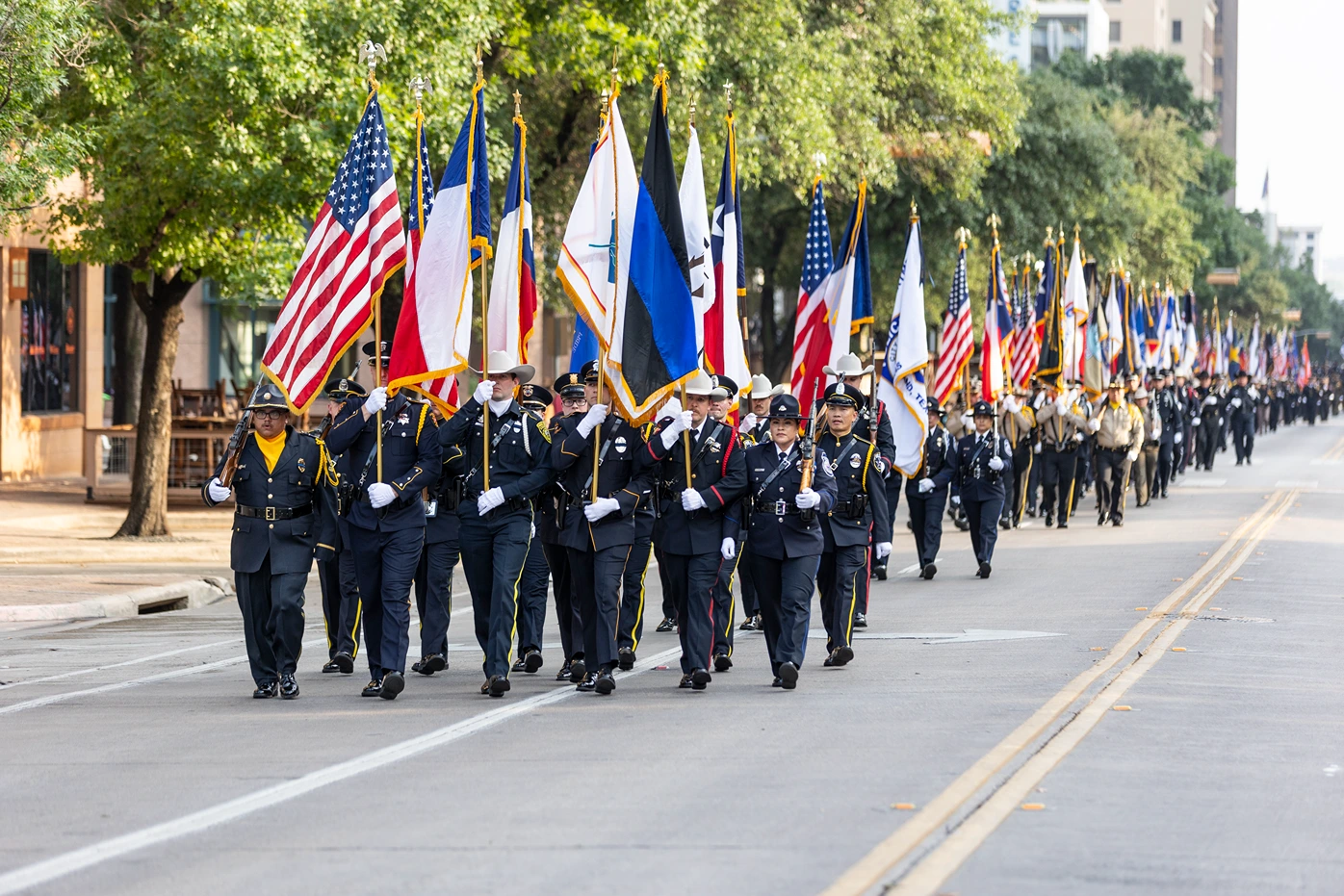 Large group of uniformed police officers marching in a street parade holding various national, state, and organizational flags.