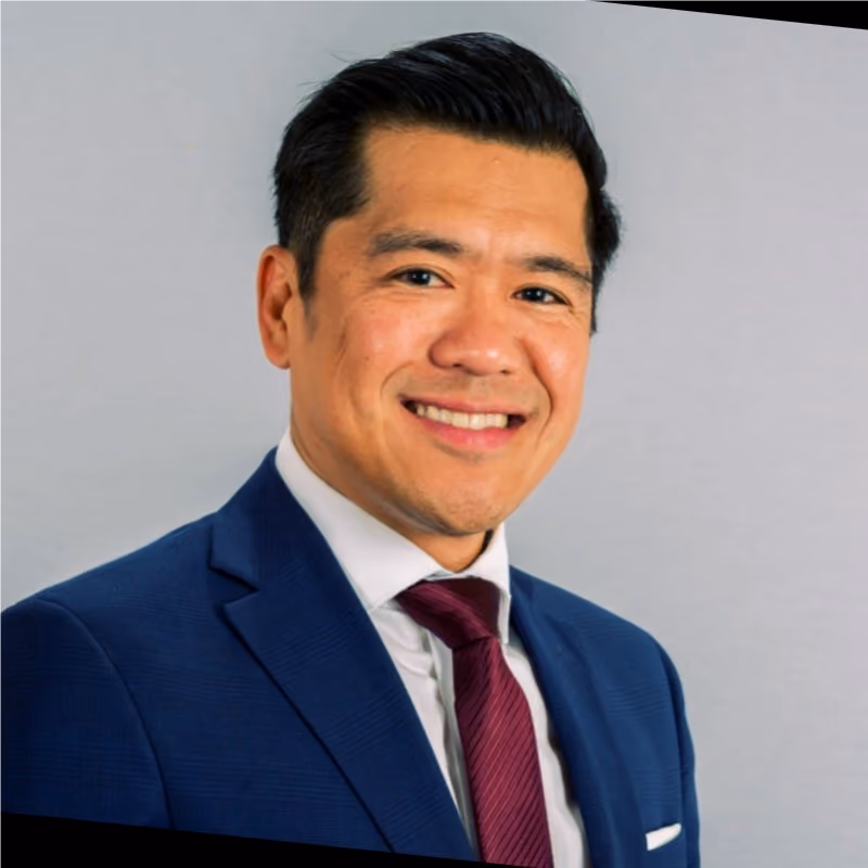 Smiling man with neatly styled black hair wearing a blue suit jacket, white dress shirt, and maroon tie, posed against a light gray background.